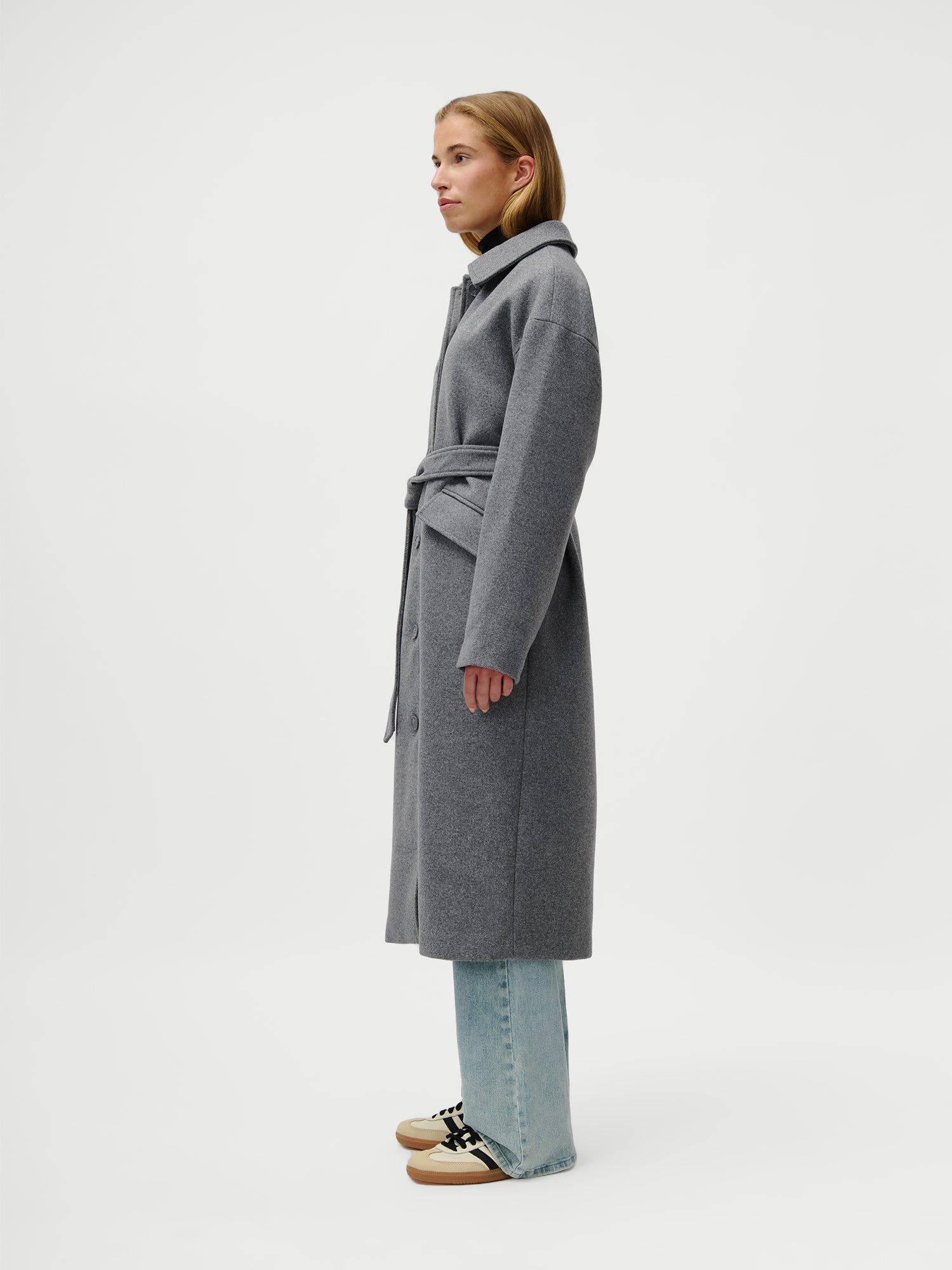 Eine Frau steht im Profil vor einem schlichten weißen Hintergrund und trägt den Anaida Coat von LeGer by Lena Gercke - einen langärmeligen, grauen Mantel mit Gürtel und lockerer Passform - über hellblauen Jeans und beigen Sneakers.