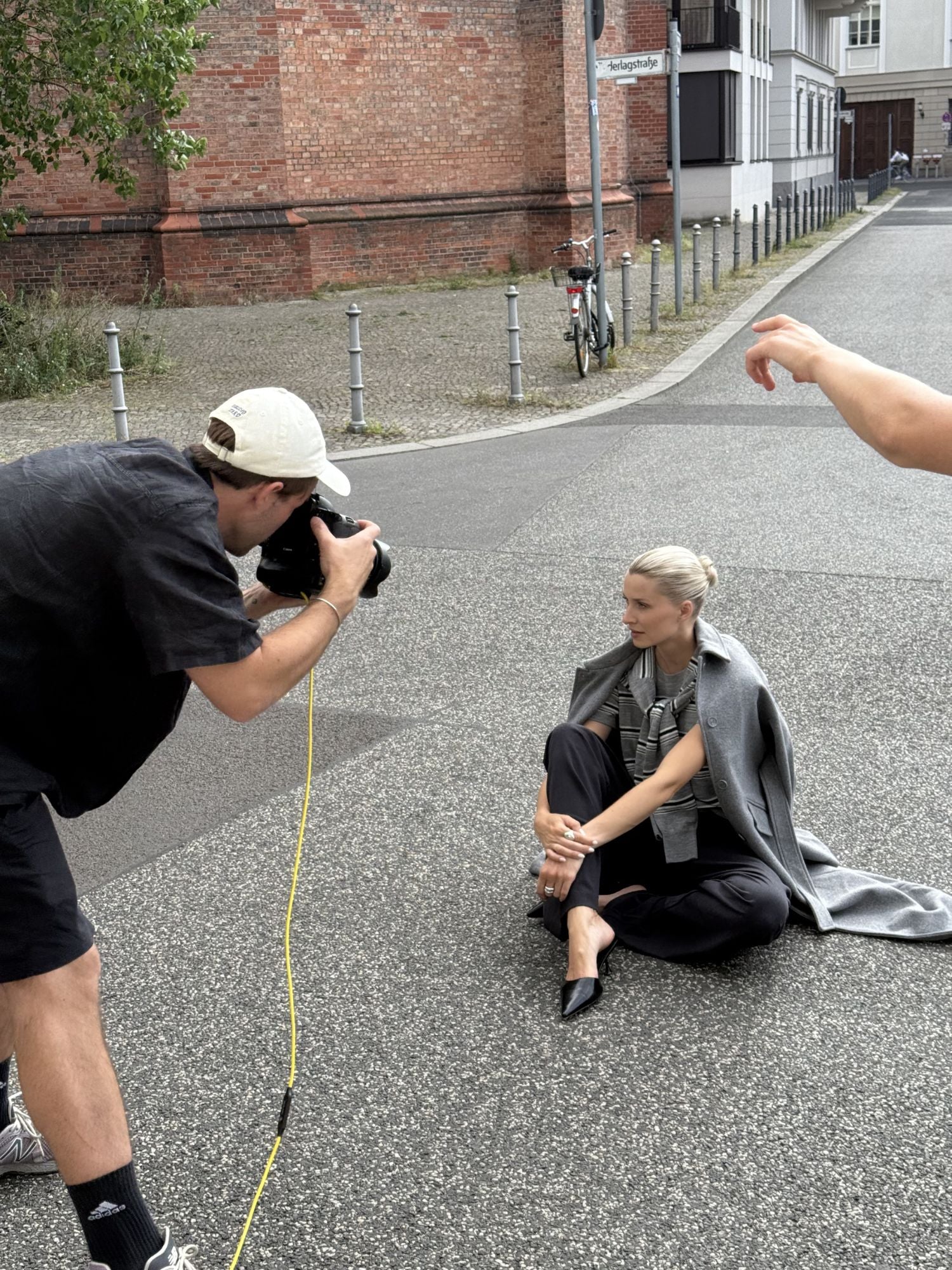 Ein Fotograf fotografiert eine Frau in einem LeGer by Lena Gercke Anaida Coat und einer schwarzen Hose auf einer Straße in der Stadt; der Arm einer anderen Person ist sichtbar, die möglicherweise hilft. In der Nähe steht ein Fahrrad.