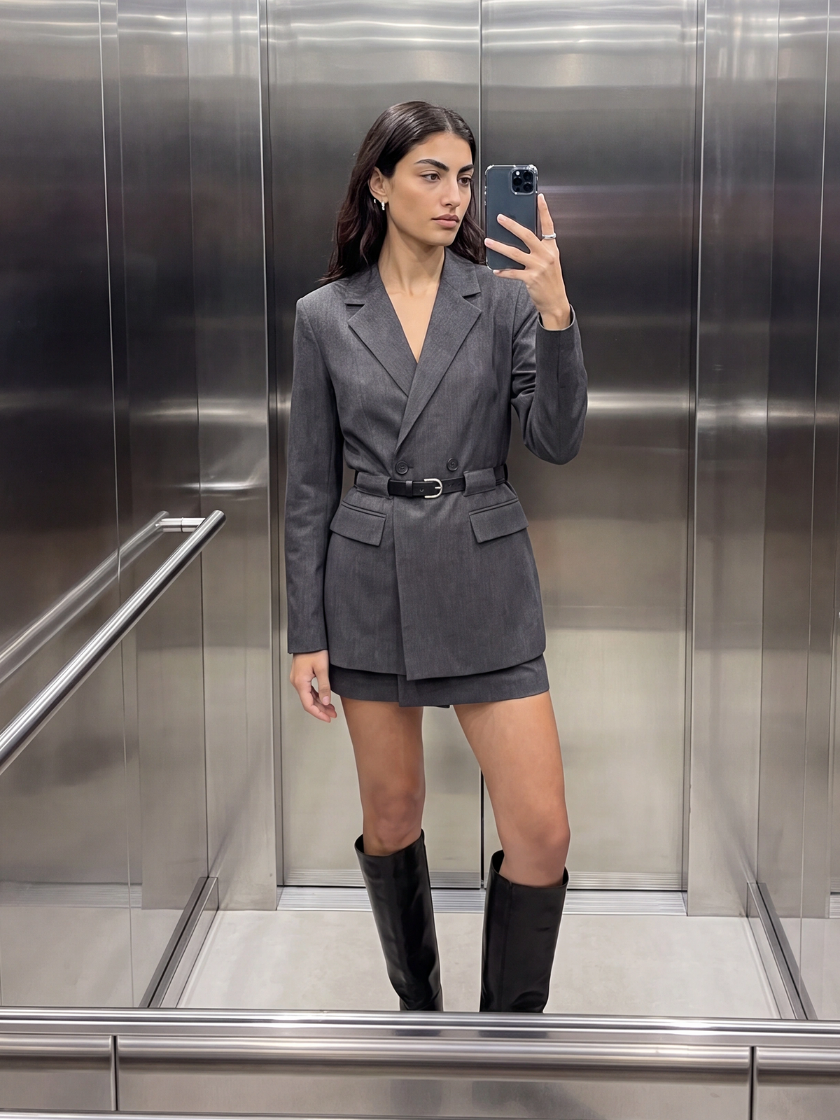 Eine Frau steht in einem Metallaufzug und macht ein Spiegel-Selfie. Sie trägt das anthrazitfarbene „Jeromina“-Kleid von LeGer by Lena Gercke mit einem Gürtel und kniehohen schwarzen Stiefeln.