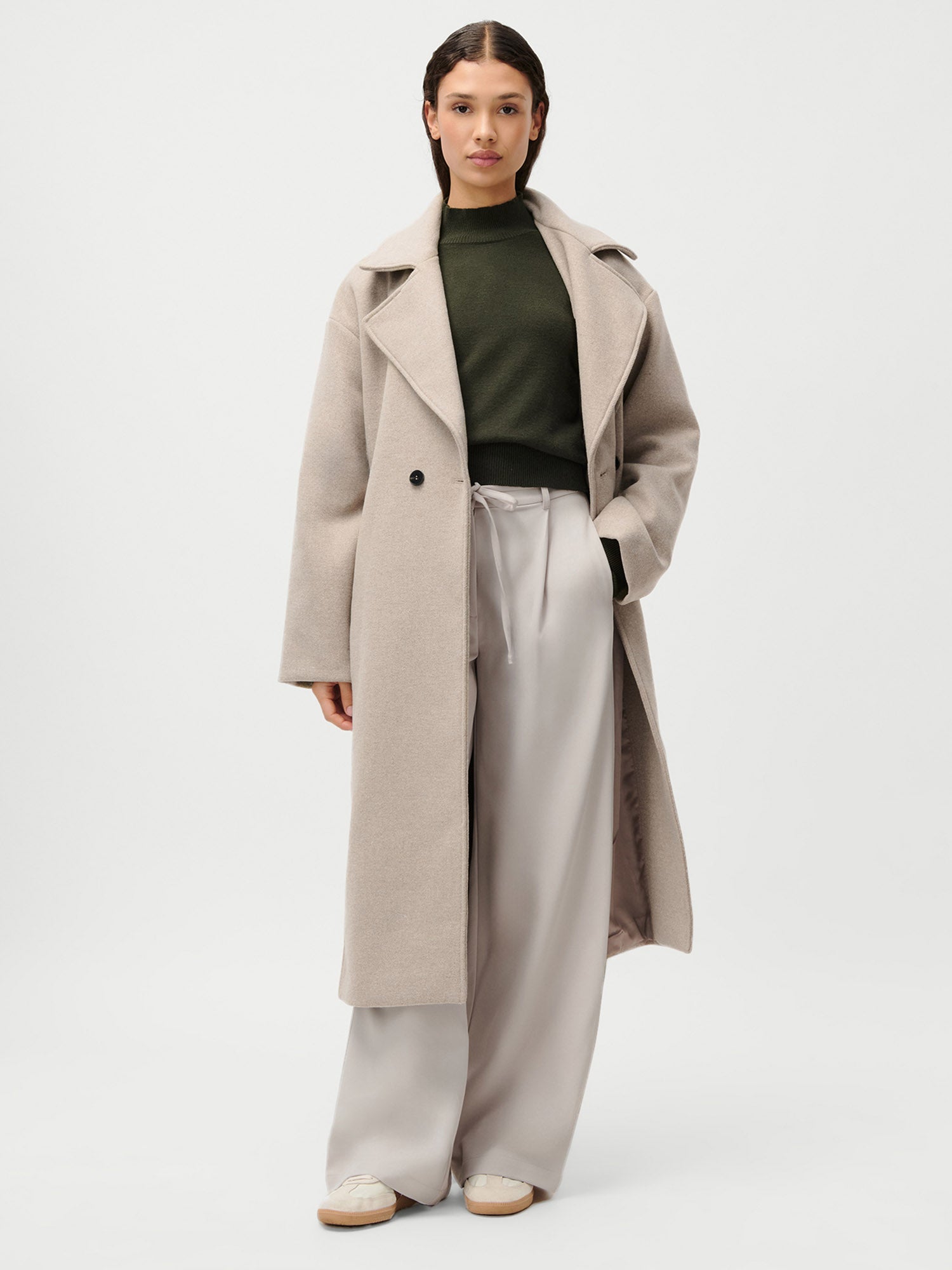 Eine Frau trägt den LeGer by Lena Gercke Nelly Mantel in Beige, gepaart mit einem olivgrünen Rollkragenpullover, einer hellgrauen Hose mit weitem Bein und weißen Sneakers. Abgebildet vor einem weißen Hintergrund in Größe 36.