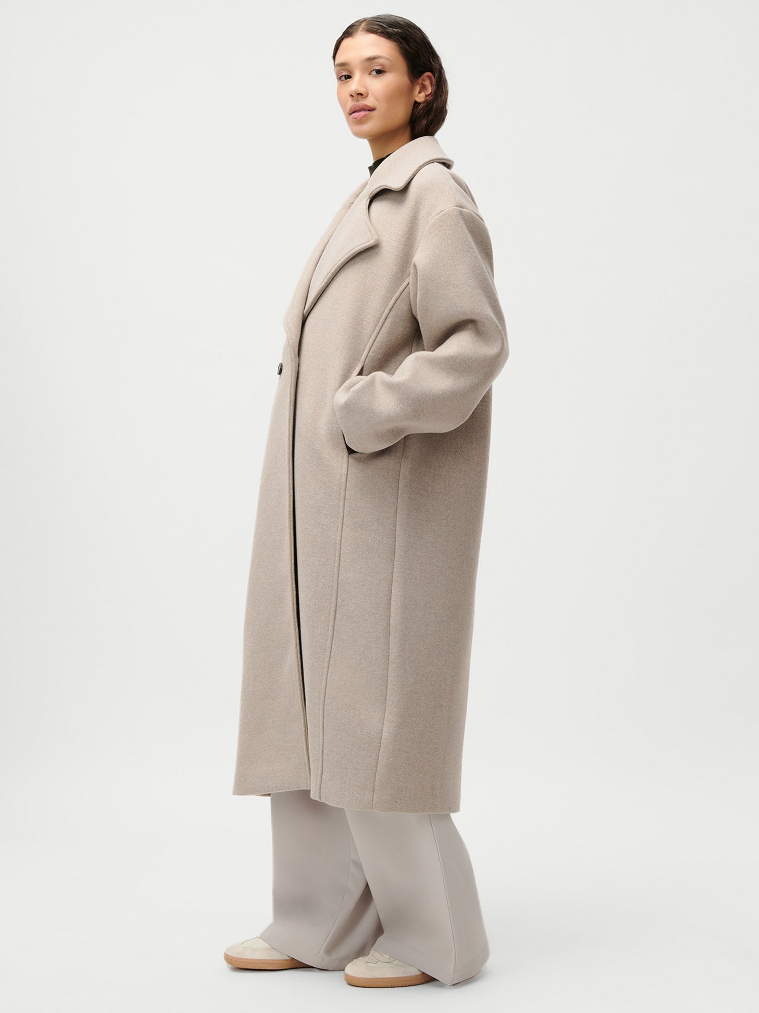 Eine Person in einem LeGer by Lena Gercke Nelly Coat, einem langen hellbeigen Mantel mit breitem Revers und normaler Passform, und einer locker sitzenden hellgrauen Hose (Größe 36), steht mit einer Hand in der Tasche vor einem schlichten hellen Hintergrund.