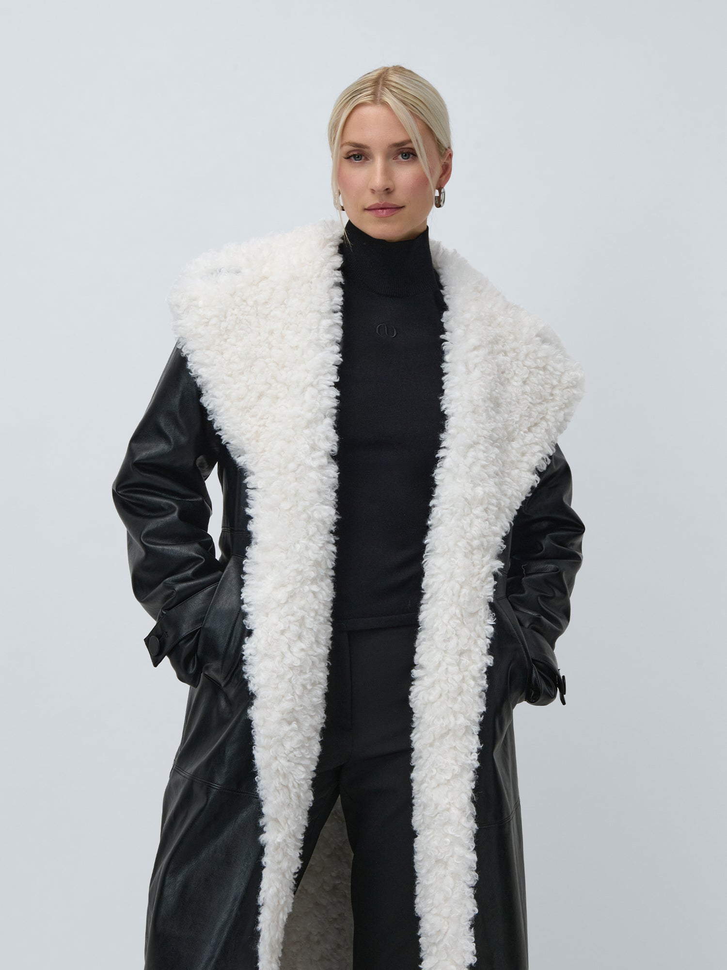 Eine blonde Person trägt den Janique Mantel von LeGer by Lena Gercke-Regular Fit mit einem großen, flauschigen weißen Shearling-Kragen und -Futter über einem schwarzen Rollkragenpullover und einer Hose vor einem schlichten hellen Hintergrund.