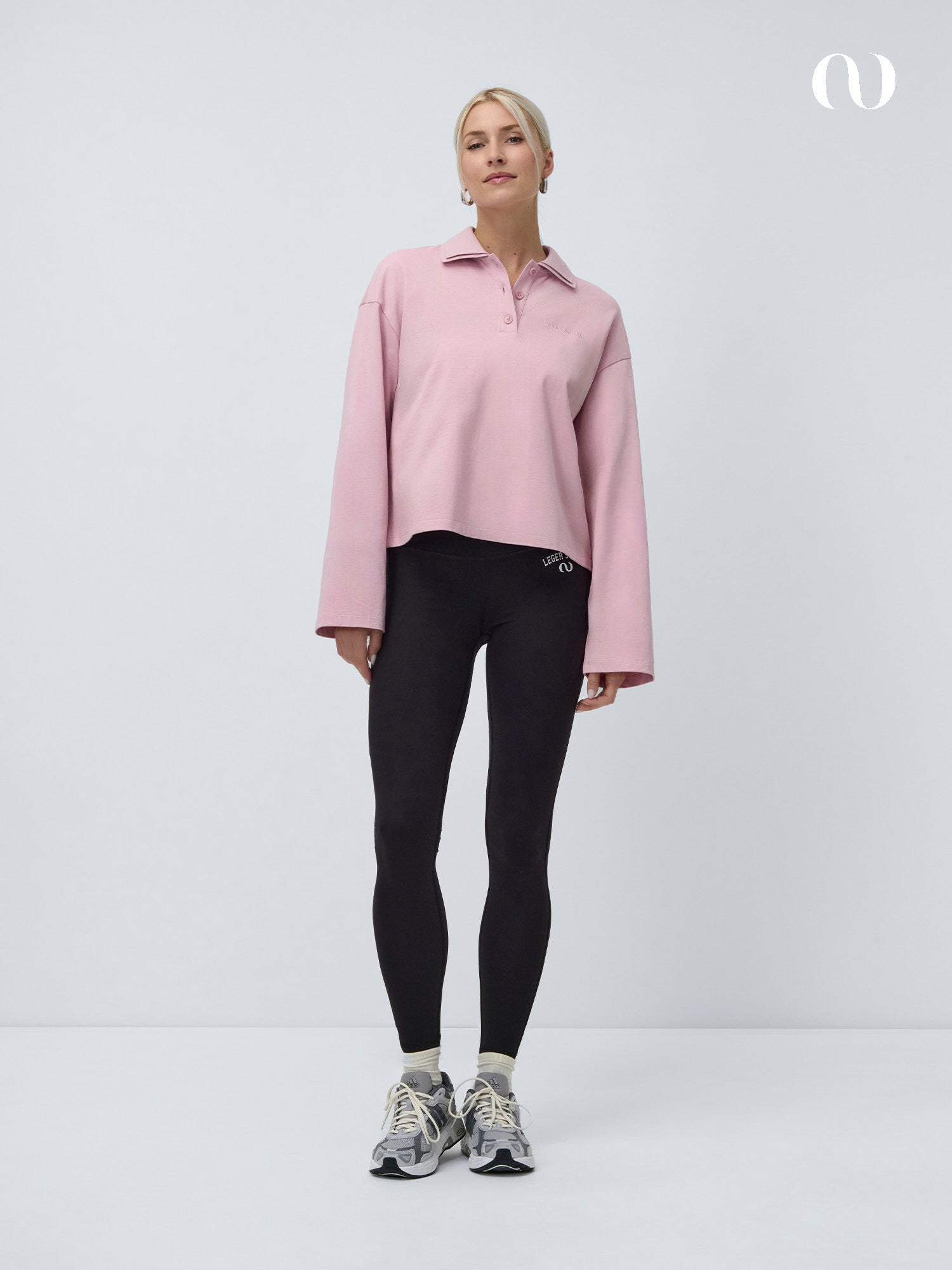 Eine Frau steht vor einem weißen Hintergrund, trägt das LeGer by Lena Gercke Pina Longsleeve in Hellrosa mit extra langen Ärmeln, gepaart mit schwarzen Leggings und grauen Turnschuhen, und schaut mit neutralem Blick nach vorne.