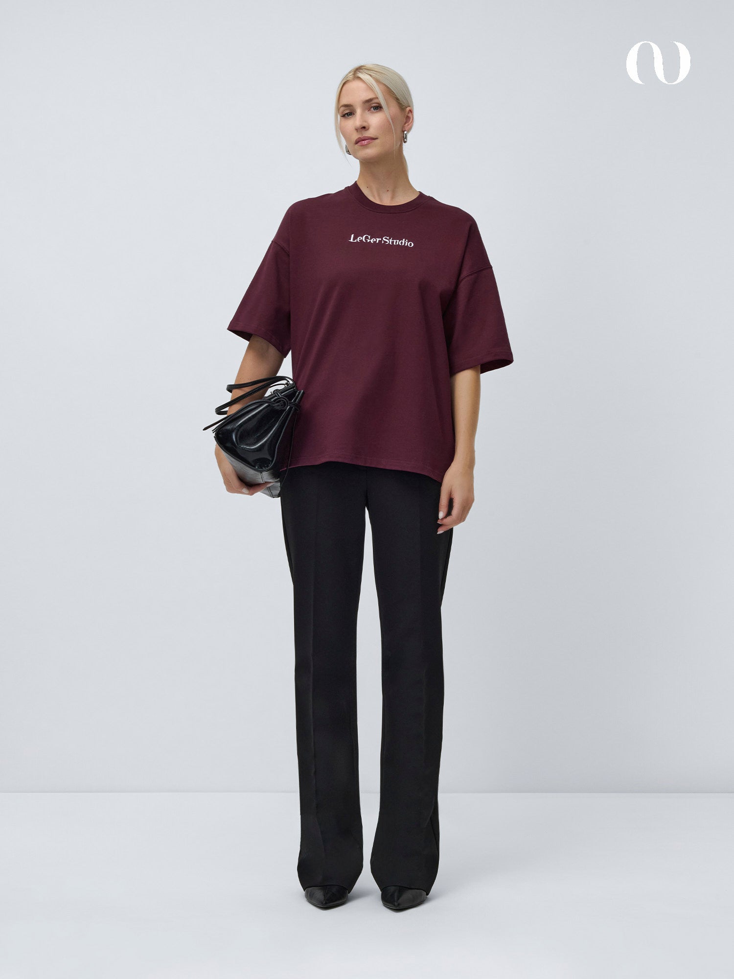 Eine Frau steht vor einem schlichten Hintergrund und trägt das Sofie T-Shirt von LeGer by Lena Gercke in kastanienbraun mit der Stickerei "Le Cour Studio", eine schwarze Hose, schwarze Schuhe und eine große schwarze Tasche. Ihr Haar ist zurückgebunden und ihr Gesichtsausdruck ist neutral.