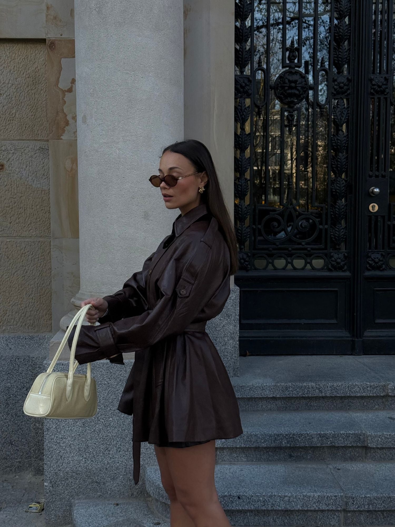 Eine Frau in der Dinorah Jacket von LeGer by Lena Gercke steht mit einer cremefarbenen Handtasche in der Hand und einer schicken Sonnenbrille auf Steinstufen vor verzierten schwarzen Türen und beigen Wänden.