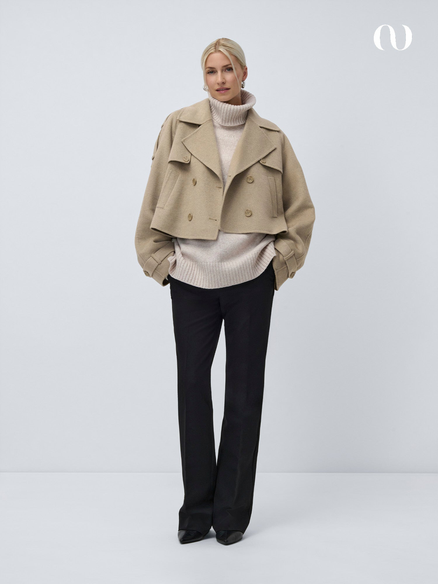 Eine Frau steht vor einem schlichten, hellen Hintergrund und trägt die Elianna Jacket von LeGer by Lena Gercke - einen beigefarbenen kurzen Trenchcoat - über einem cremefarbenen Rollkragenpullover, einer schwarzen maßgeschneiderten Hose und schwarzen spitzen Schuhen.