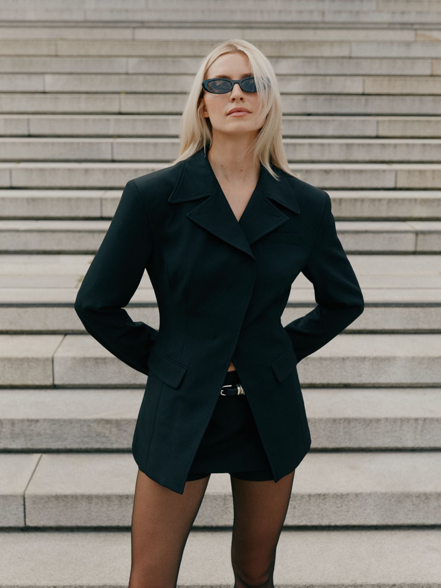 Eine Frau mit langen blonden Haaren, dunkler Sonnenbrille und dem Ismey Blazer von LeGer by Lena Gercke, gestylt mit schwarzen Shorts und durchsichtigen Strumpfhosen, steht selbstbewusst auf steinernen Stufen, die Hände in die Hüften gestemmt.