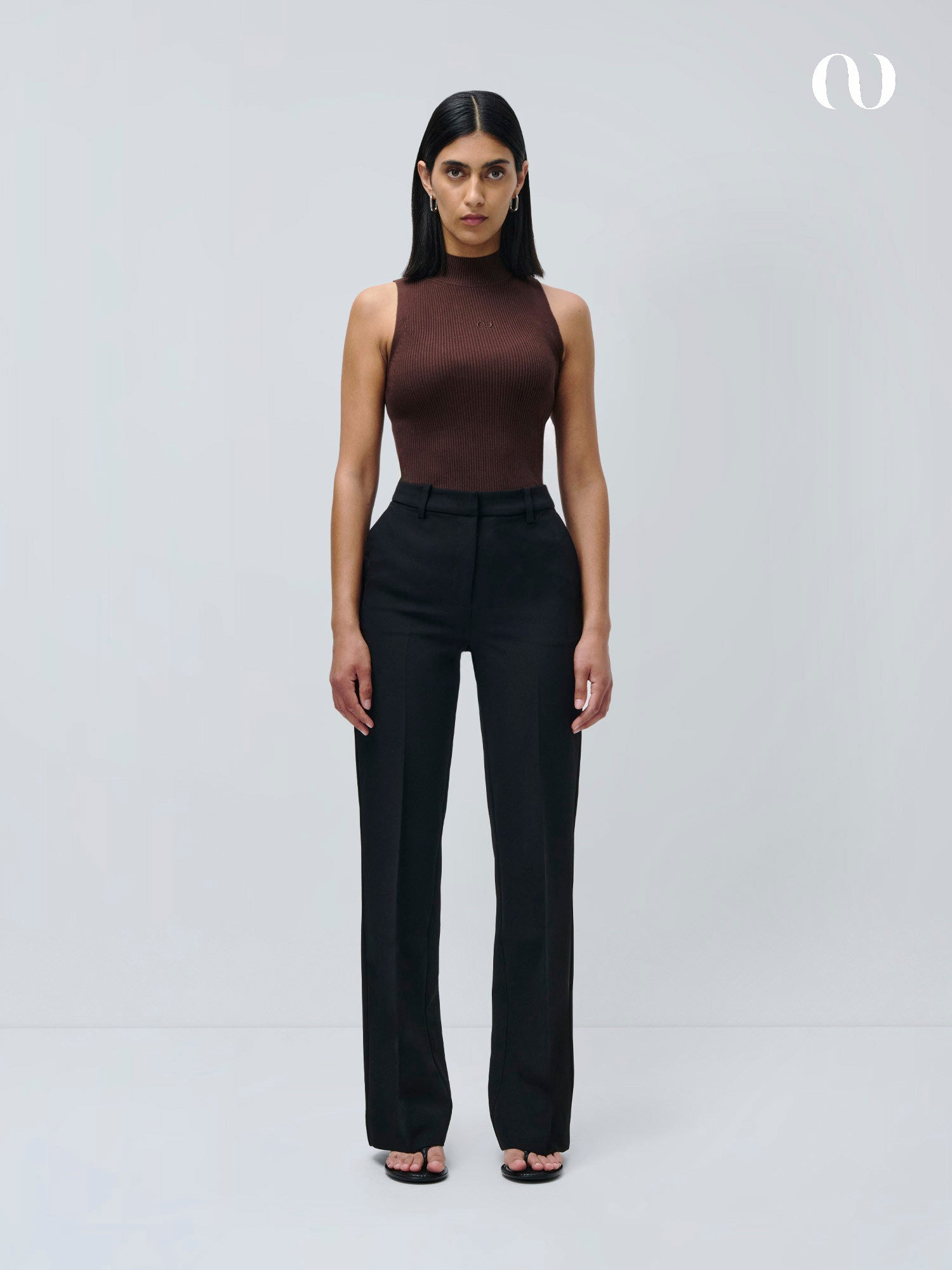 Eine Frau mit glattem dunklem Haar steht vor einem schlichten hellgrauen Hintergrund und modelliert die Fatou Pants von LeGer by Lena Gercke in schmaler Passform und langer Länge. Sie trägt ein ärmelloses braunes Oberteil und schwarze, offene Absätze und blickt mit einem neutralen Gesichtsausdruck nach vorne.