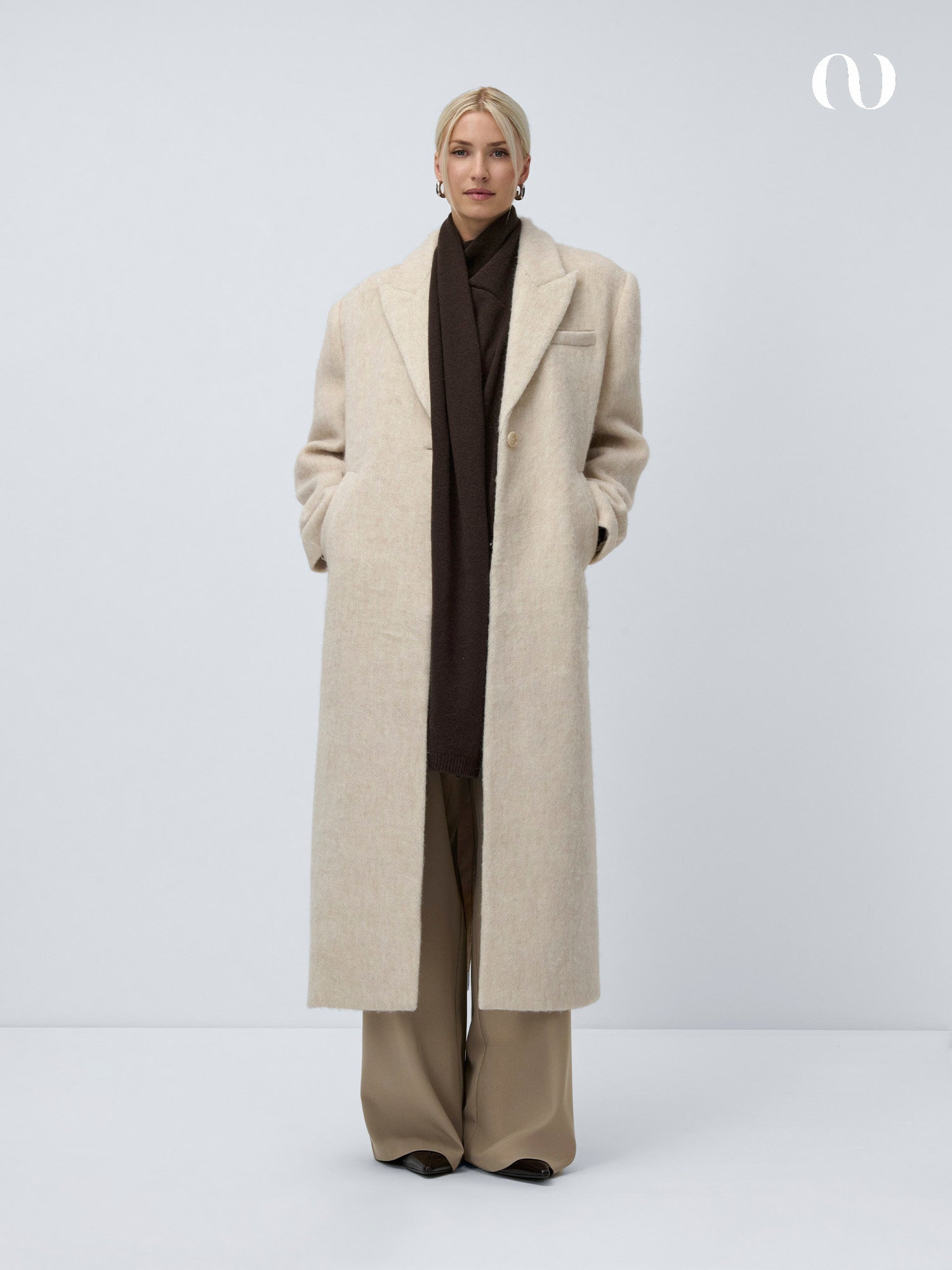 Eine Person mit hellem Haar trägt den cremefarbenen Helga-Pullover von LeGer by Lena Gercke über einer beigefarbenen Hose mit weiten Beinen und einem dunkelbraunen Schal. Sie steht vor einem schlichten hellen Hintergrund und hat die Hände in den Manteltaschen.