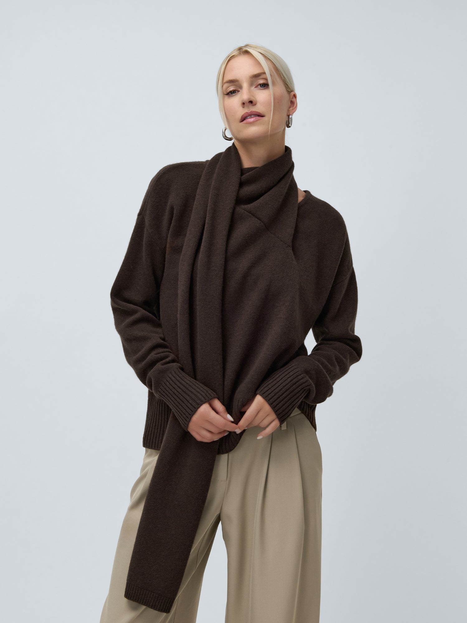 Eine blonde Frau trägt den Helga-Pullover von LeGer by Lena Gercke mit einem passenden langen Schal und einer beigen Hose mit weitem Bein vor einem schlichten hellgrauen Hintergrund.