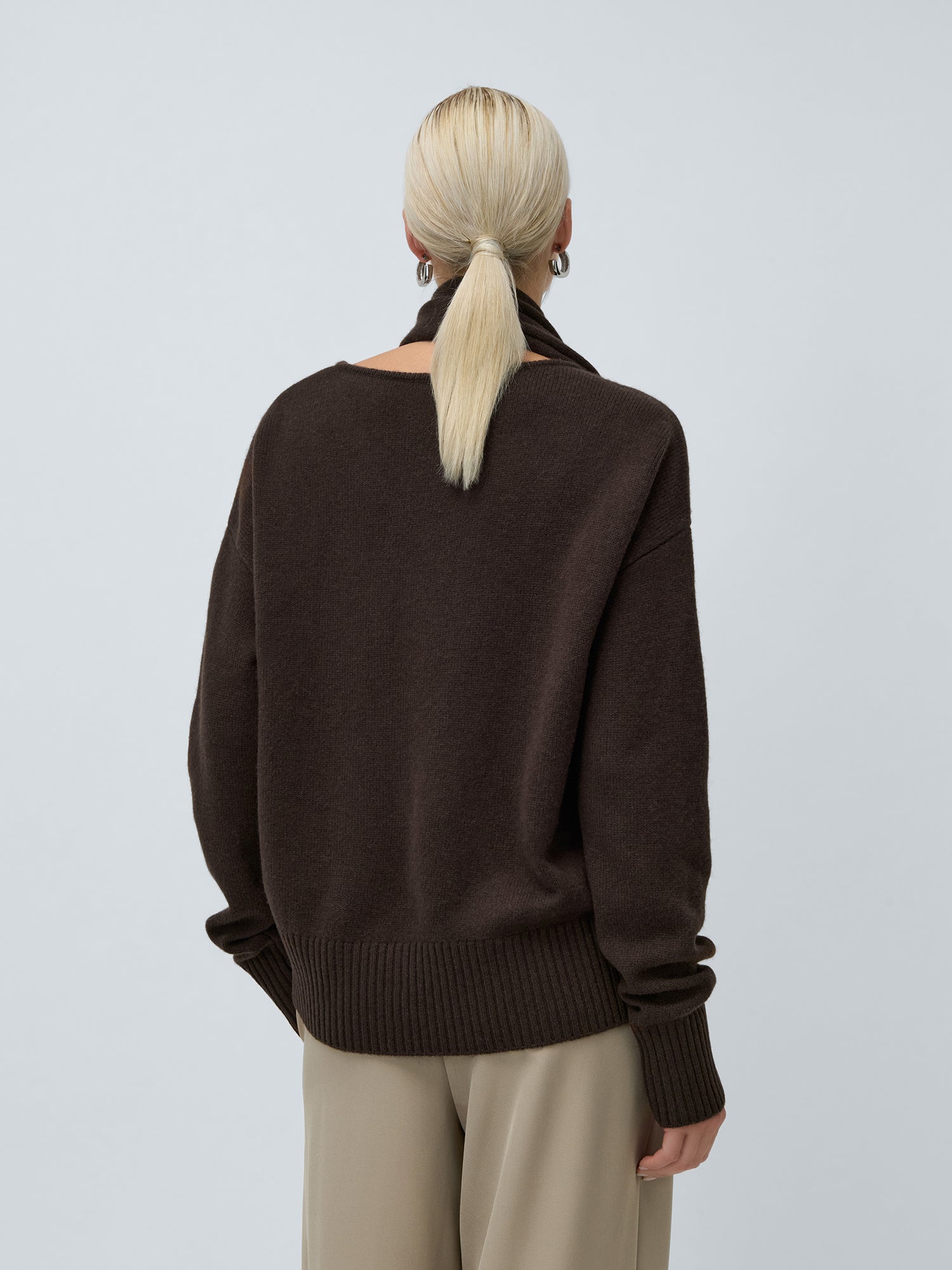 Eine Person mit glattem blondem Haar in einem niedrigen Pferdeschwanz ist von hinten zu sehen. Sie trägt den LeGer by Lena Gercke Helga Jumper in Dunkelbraun und eine beigefarbene Hose vor einem schlichten hellgrauen Hintergrund.