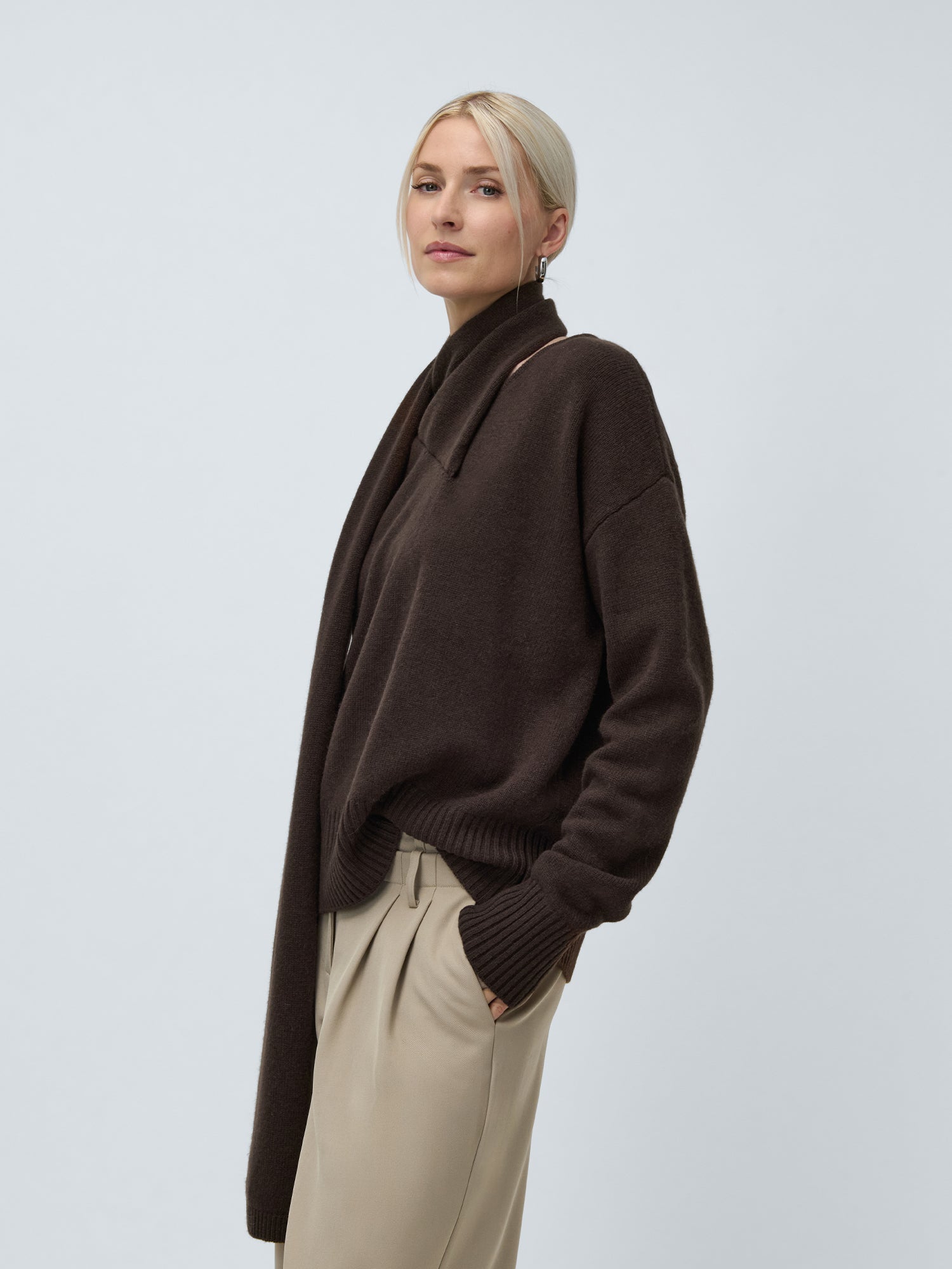 Eine blonde Frau mit tiefem Dutt trägt den Helga Jumper von LeGer by Lena Gercke - einen dunkelbraunen, locker sitzenden Pullover - zusammen mit einem passenden Schal und einer beigen Hose und steht mit einer Hand in der Tasche vor einem hellgrauen Hintergrund.