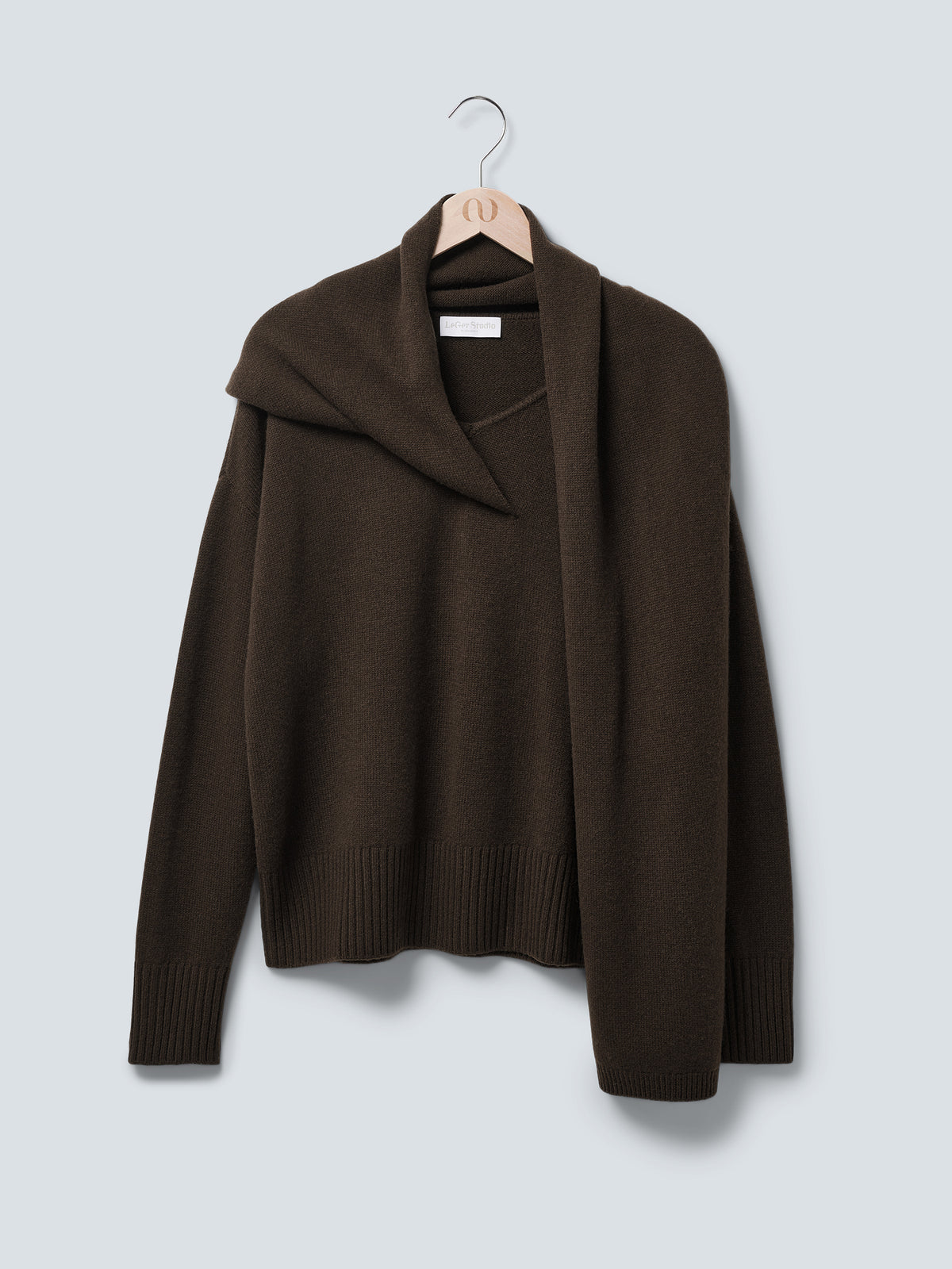 Der Pullover „Helga“ von LeGer by Lena Gercke ist ein dunkelbrauner, locker sitzender, langer Pullover mit einem passenden Schal, der über eine Schulter drapiert ist. Er wird auf einem Holzkleiderbügel vor einem hellgrauen Hintergrund präsentiert.