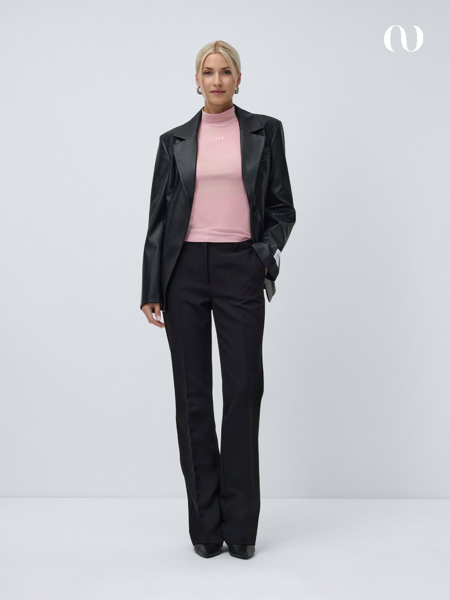 Eine Frau mit zurückgebundenen blonden Haaren steht vor einem weißen Hintergrund und trägt ein rosa LeGer by Lena Gercke Franka Longsleeve-Top, einen schwarzen Lederblazer, eine schmal geschnittene schwarze Hose und schwarze Schuhe, wobei sie eine Hand in der Tasche hat.