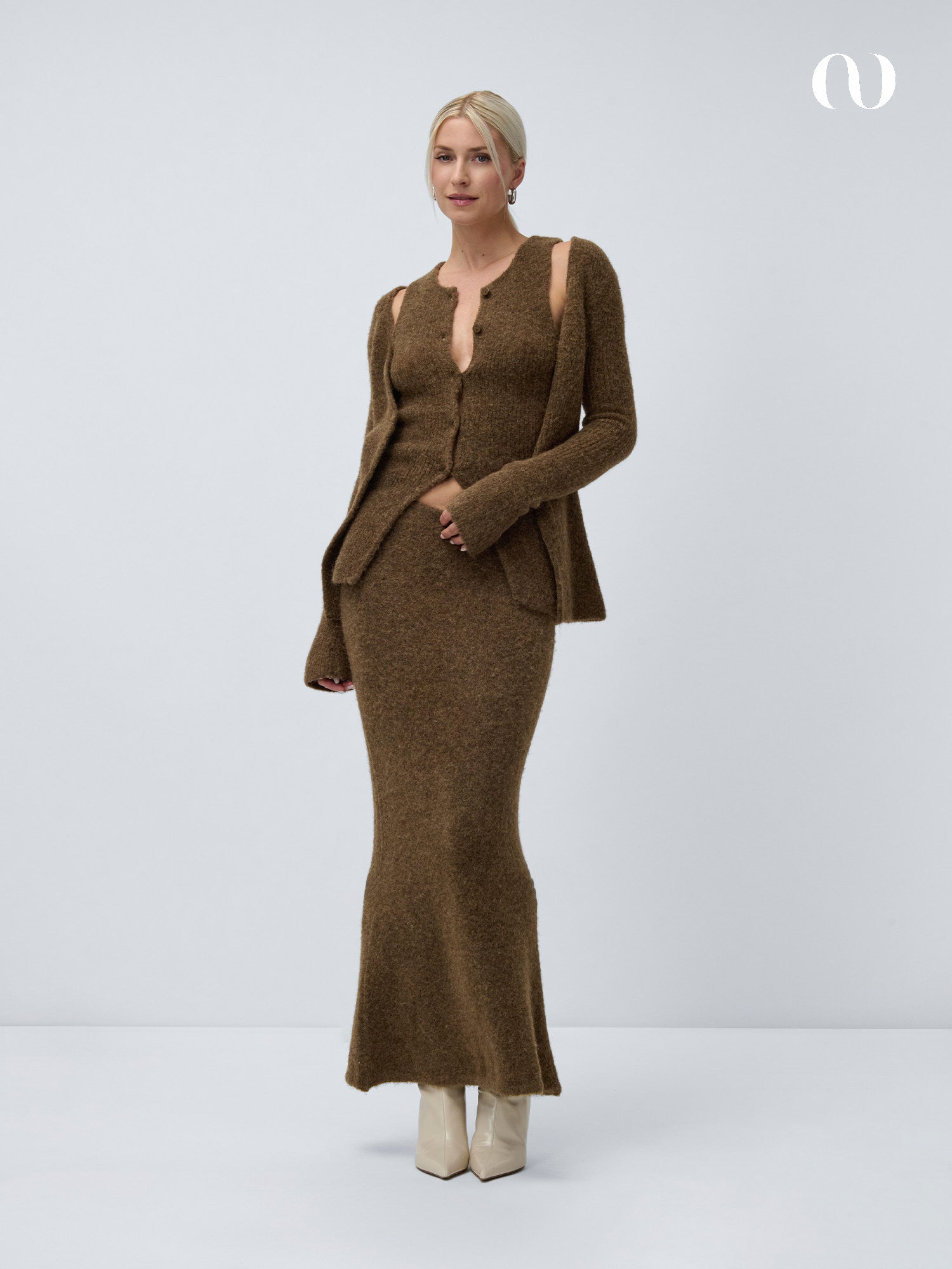 Eine Frau mit blondem, hochgestecktem Haar trägt einen braunen, strukturierten Slim Fit Zweiteiler mit dem LeGer by Lena Gercke Frederike Top und einem passenden, taillierten Maxirock, gepaart mit weißen Stiefeln, vor einem schlichten, hellgrauen Hintergrund.