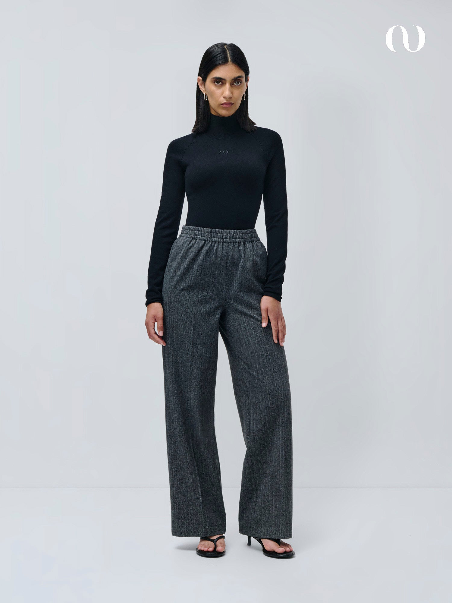 Eine Frau steht vor einem hellgrauen Hintergrund und trägt einen schwarzen Rollkragenpullover, LeGer by Lena Gercke Shannon Pants mit weiten Beinen und einer lockeren Passform, dazu schwarze Sandalen mit Absatz. Ihr glattes Haar ist in der Mitte gescheitelt.