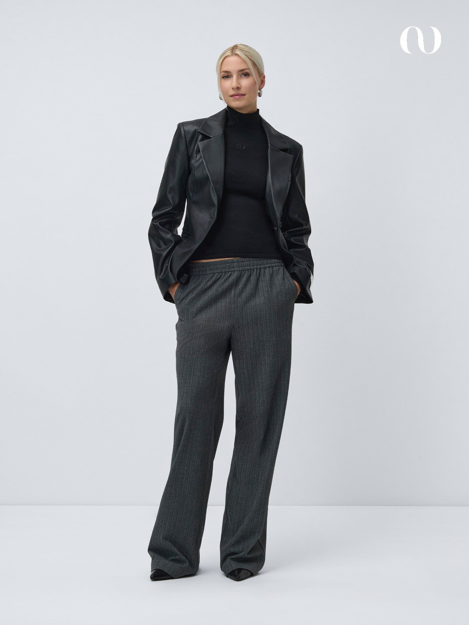 Eine Frau mit blondem, nach hinten gestyltem Haar steht vor einem weißen Hintergrund und trägt einen schwarzen Rollkragenpullover, einen schwarzen Lederblazer und die LeGer by Lena Gercke Shannon Pants Tall in Grau mit weitem Bein und Maxi-Länge, die Hände in den Taschen.