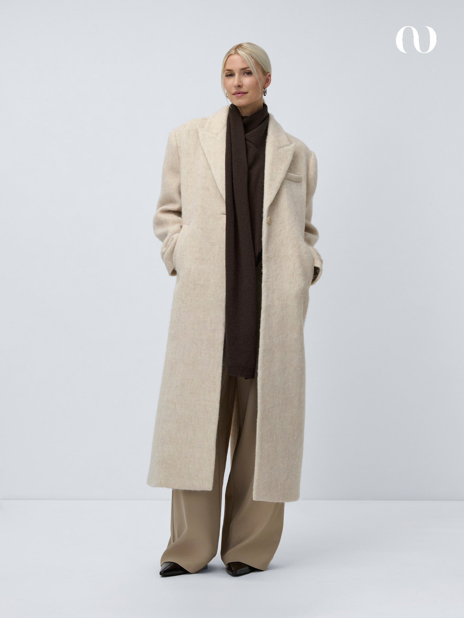 Eine Person steht vor einem schlichten, hellen Hintergrund und trägt einen langen, hellbeigen Mantel über einem dunkelbraunen Rollkragenpullover und LeGer by Lena Gercke Marcelina Pants Tall mit weiten Beinen, die Hände in den Manteltaschen. Der Look ist minimalistisch und elegant.
