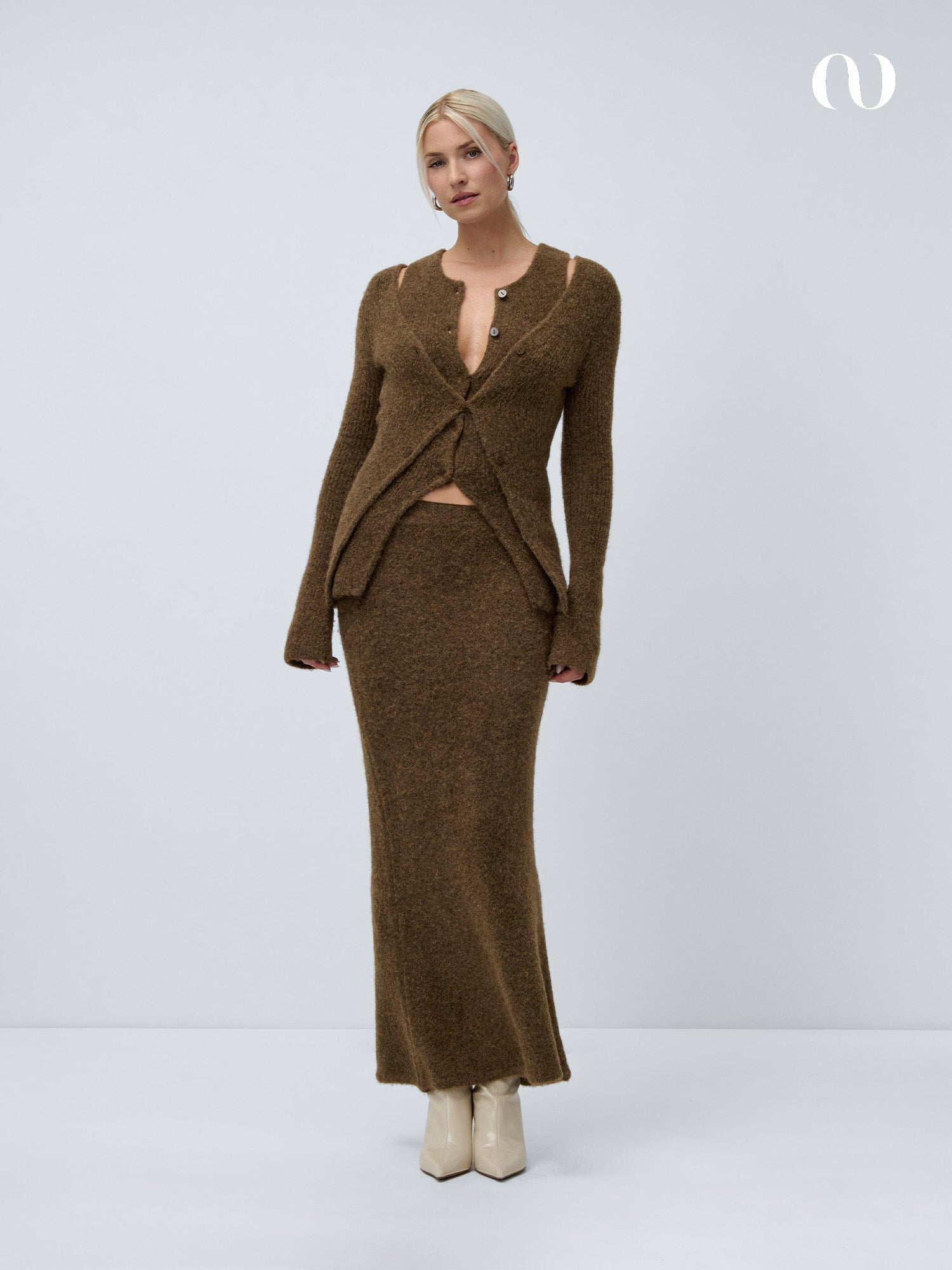 Eine Frau posiert vor einem hellgrauen Hintergrund in einem schmal geschnittenen Frederike Cardigan von LeGer by Lena Gercke mit tiefem V-Ausschnitt und einem dazu passenden langen Rock. Ihr helles Haar ist zu einer niedrigen Hochsteckfrisur gestylt und wird durch cremefarbene Stiefel mit Absatz ergänzt.