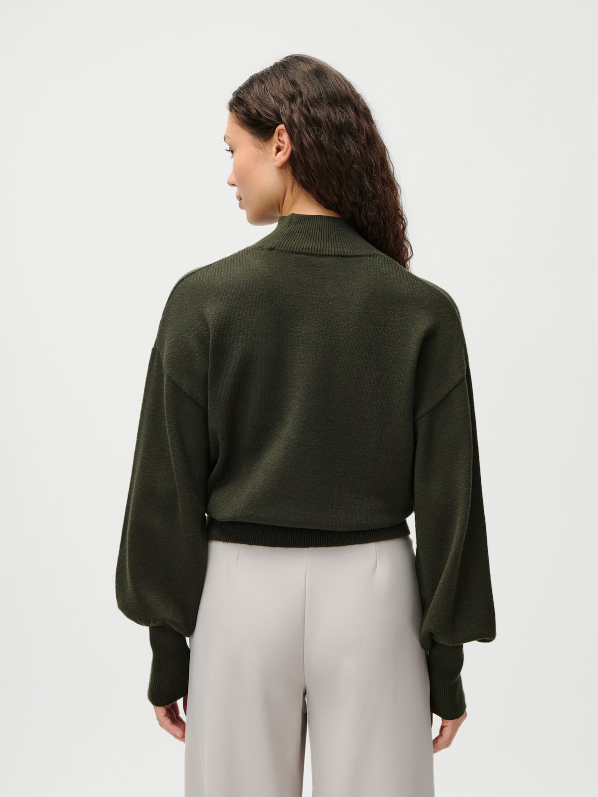 Eine Frau mit langen, welligen braunen Haaren steht mit dem Rücken zur Kamera und trägt den Pullover „Penelope“ von LeGer by Lena Gercke in Olivgrün mit Ballonärmeln und einem dezenten Stehkragen, dazu eine hellbeige Hose mit hoher Taille vor einem schlichten hellen Hintergrund.