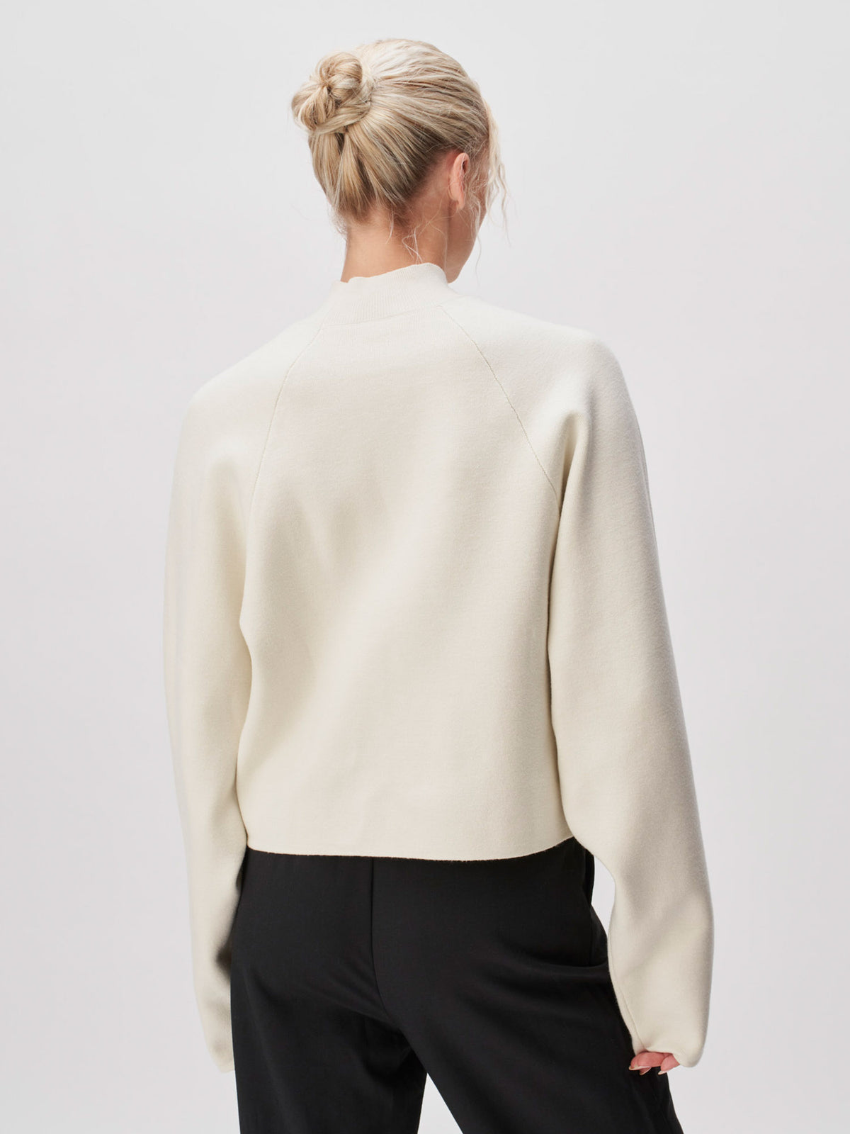 Eine Person mit blonden Haaren, die zu einem Dutt zusammengebunden sind, trägt den cremefarbenen Pullover „Heather“ von LeGer by Lena Gercke – mit langen Ärmeln und normaler Länge –, dazu eine schwarze Hose, und steht mit dem Rücken zum Betrachter vor einem schlichten, hellen Hintergrund.