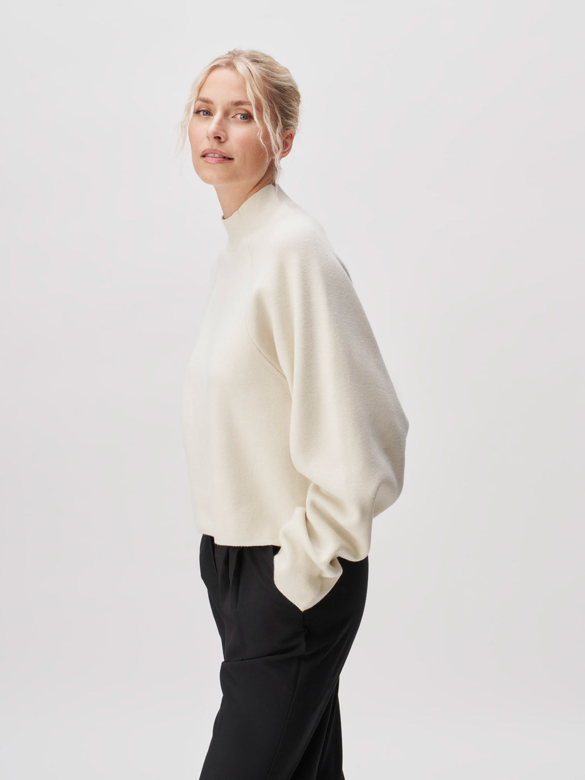 Eine Person mit blonden Haaren, die zu einem lockeren Dutt zusammengebunden sind, trägt den cremefarbenen Pullover „Heather“ von LeGer by Lena Gercke in normaler Passform sowie eine schwarze Hose. Sie steht vor einem schlichten, hellen Hintergrund und blickt leicht über die Schulter.