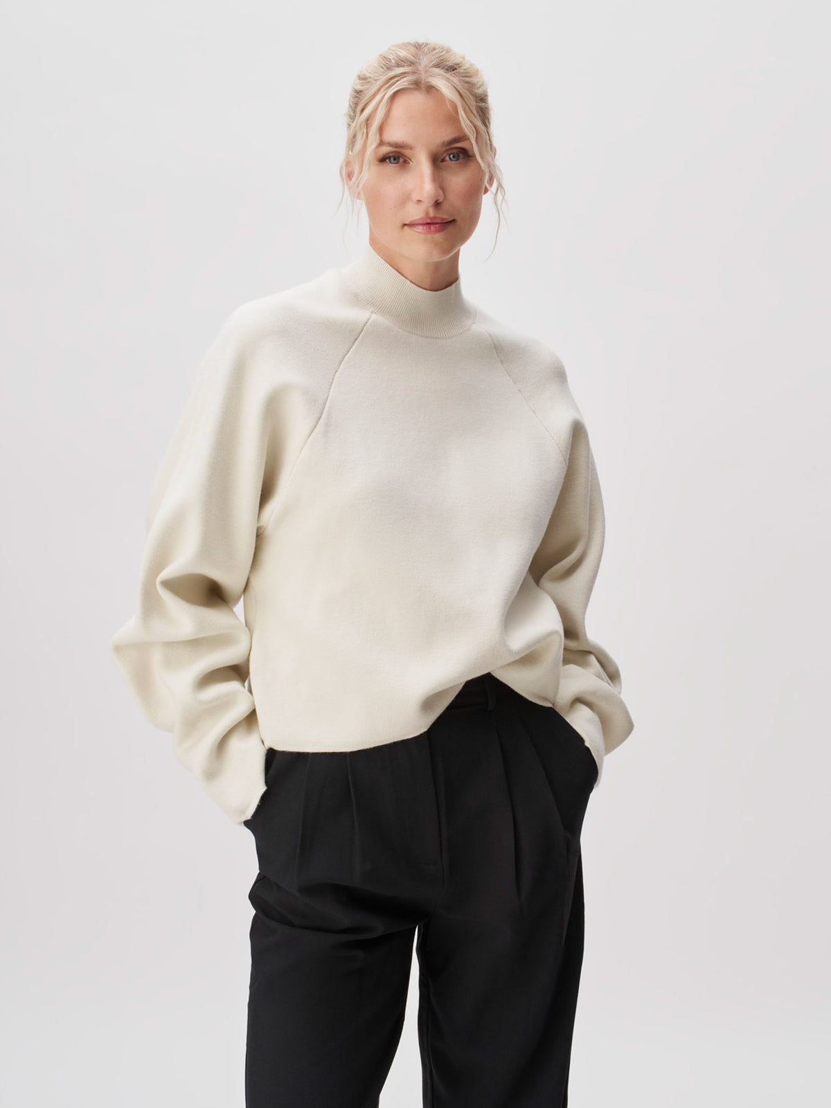 Eine Person mit blonden Haaren, die zu einem lockeren Dutt zusammengebunden sind, trägt den Pullover „Heather“ von LeGer by Lena Gercke – einen cremefarbenen Langarmpullover mit hohem Kragen und normaler Länge –, kombiniert mit einer schwarzen Hose, und steht mit den Händen in den Taschen vor einem schlichten, hellen Hintergrund.