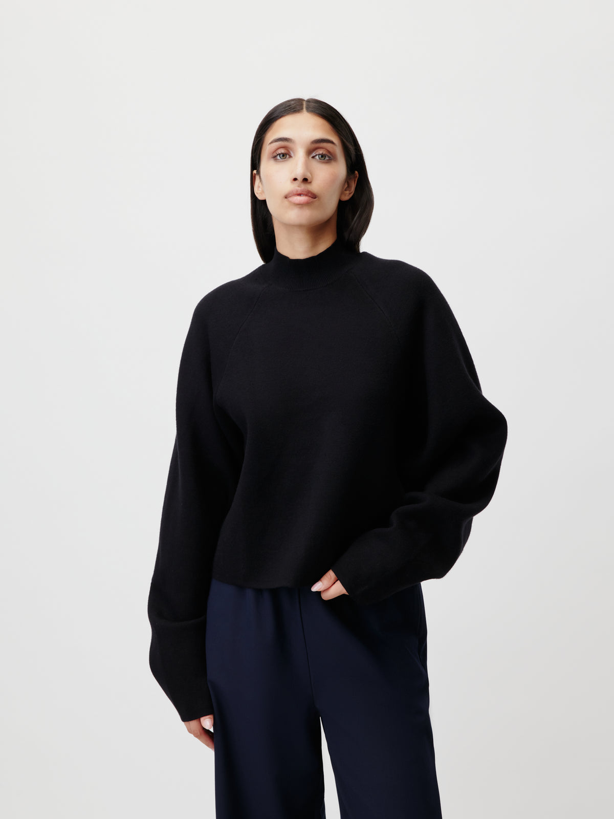 Eine Person mit glattem, schulterlangem, dunklem Haar trägt den Pullover „Heather“ von LeGer by Lena Gercke mit Stehkragen und Raglanärmeln sowie eine dunkle Hose und steht vor einem schlichten hellgrauen Hintergrund.
