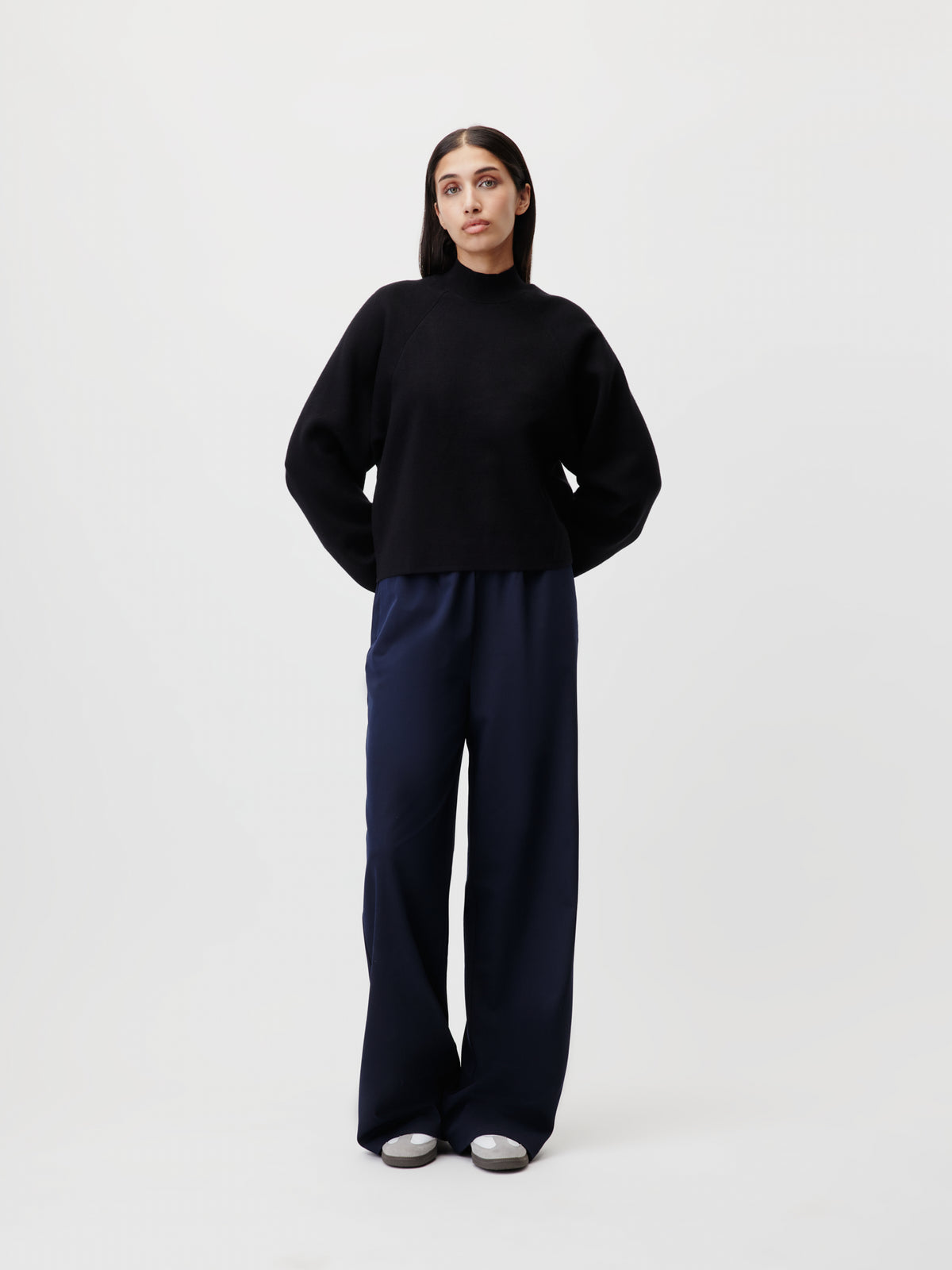 Eine Person steht vor einem schlichten weißen Hintergrund und trägt den „Heather“-Pullover von LeGer by Lena Gercke – einen locker geschnittenen schwarzen Pullover mit Stehkragen und Raglanärmeln. Dazu trägt sie weite marineblaue Hosen und silberne Schuhe. Die Hände sind hinter dem Rücken verschränkt, der Blick ist leicht zur Seite gerichtet.
