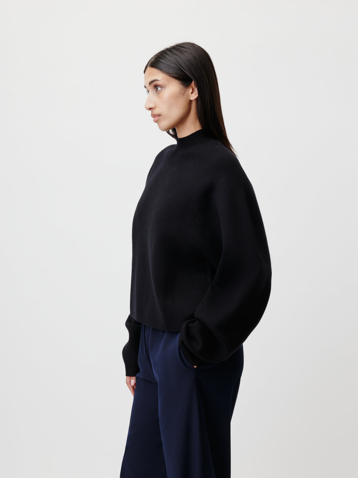 Eine Frau mit langen dunklen Haaren, gekleidet in den Pullover „Heather“ von LeGer by Lena Gercke – einen schwarzen Pullover mit Raglanärmeln und Stehkragen –, dazu eine marineblaue Hose, steht im Profil vor einem schlichten weißen Hintergrund.