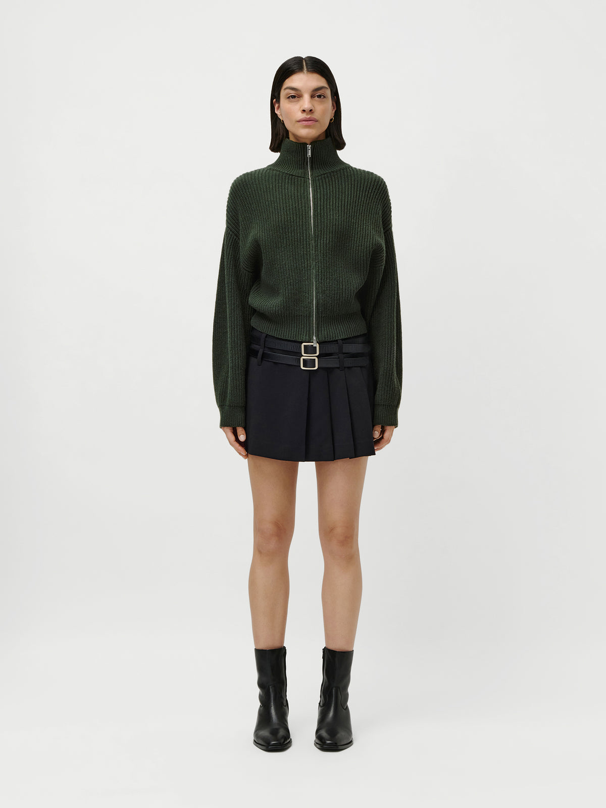 Eine Person steht mit dem Gesicht nach vorne vor einem schlichten weißen Hintergrund und trägt einen dunkelgrünen „Cindy“-Pullover von LeGer by Lena Gercke mit Reißverschluss vorne und normaler Länge, einen kurzen schwarzen Faltenrock mit einem Gürtel mit zwei Schnallen in Größe 36 sowie schwarze Stiefeletten.