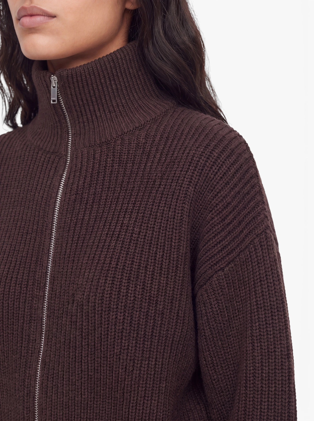 Eine Frau, die den Pullover „Cindy“ von LeGer by Lena Gercke in dunkelbraunem Rippstrick mit Reißverschluss trägt, gezeigt von den Schultern aufwärts, mit langen, gewellten Haaren und vor einem neutralen Hintergrund.