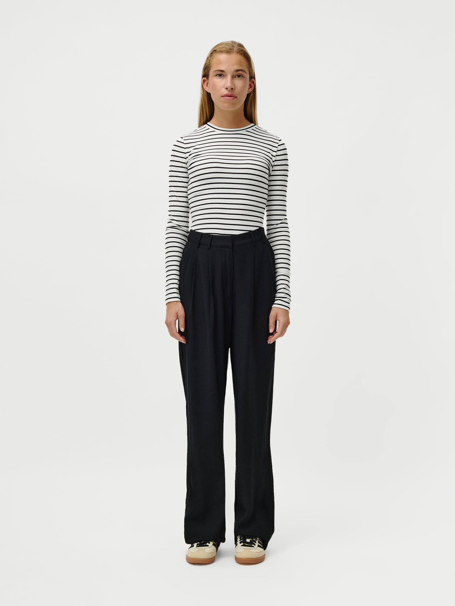 Eine junge Frau steht mit dem Gesicht nach vorne vor einem schlichten weißen Hintergrund und trägt das schwarz-weiß gestreifte Cinzia Longsleeve von LeGer by Lena Gercke, eine schwarze Hose mit hoher Taille in normaler Länge und beigefarbene Turnschuhe.