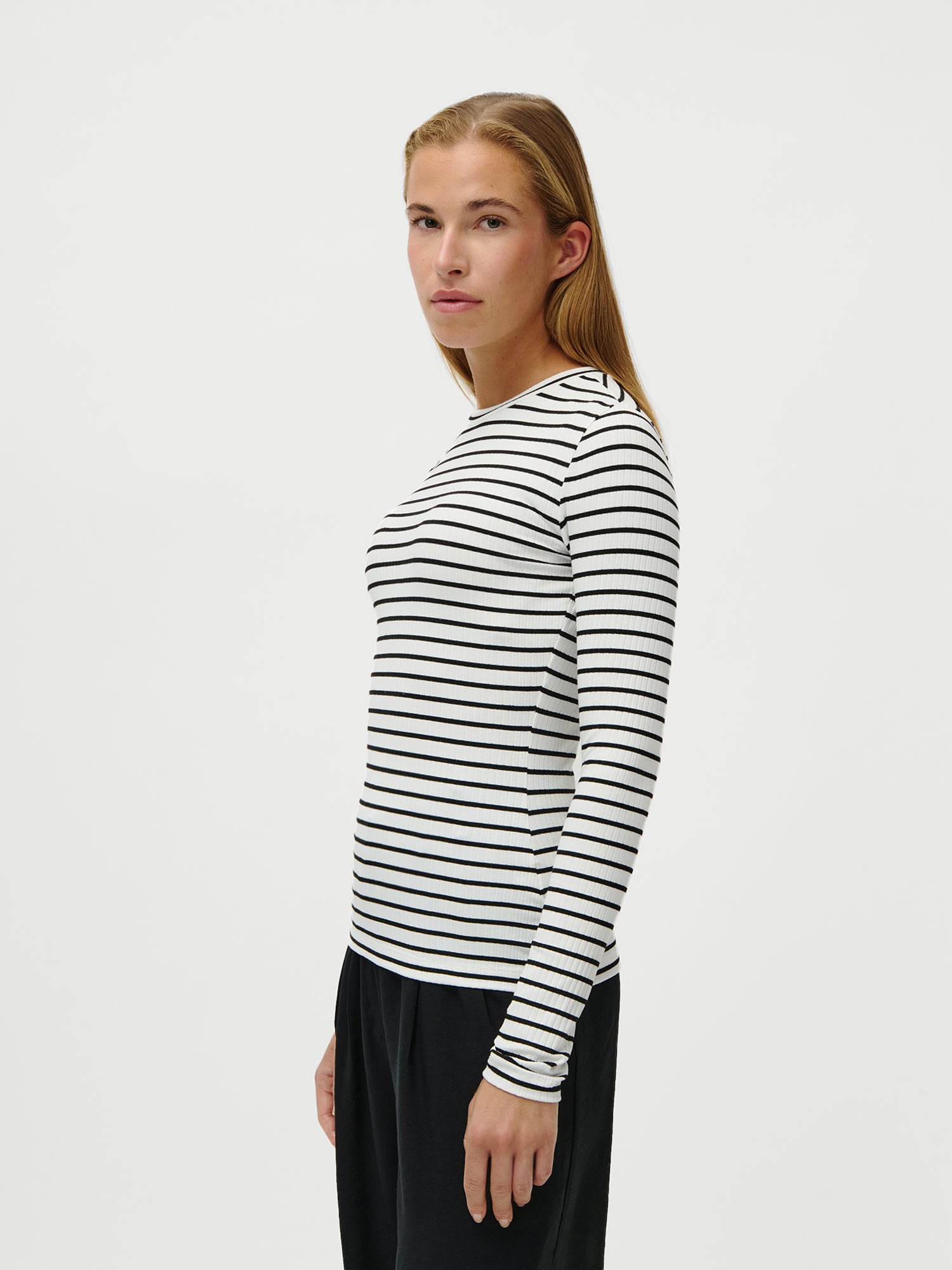 Eine Person mit langem, glattem, hellbraunem Haar steht seitlich und trägt das LeGer by Lena Gercke Cinzia Longsleeve - ein schwarz-weiß gestreiftes Hemd in normaler Länge - und eine schwarze Hose vor einem schlichten weißen Hintergrund.