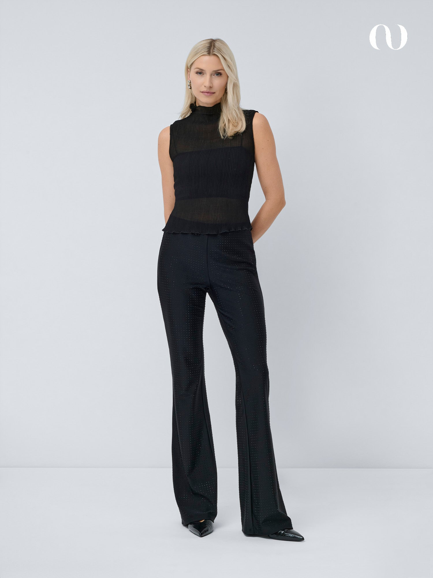 Eine Frau mit glattem blondem Haar trägt ein ärmelloses schwarzes Oberteil, schwarze spitze Schuhe und die Daniela-Hose von LeGer by Lena Gercke in normaler Passform mit weitem Bein und dezentem Glitzern, die vor einem schlichten hellgrauen Hintergrund steht.