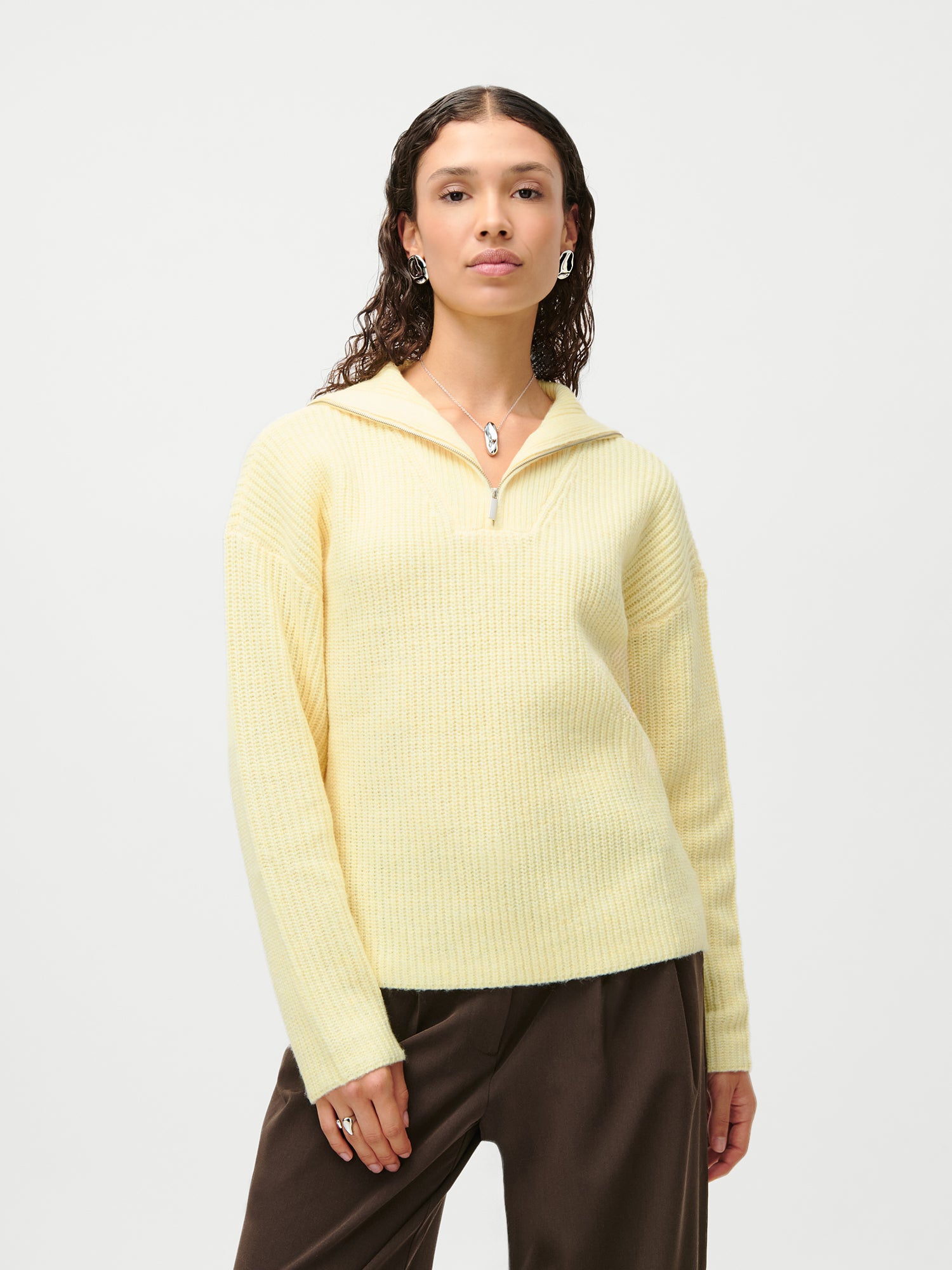 Eine Person mit gewellten dunklen Haaren trägt den LeGer x Purelei - Lea Jumper von LeGer by Lena Gercke in Hellgelb mit einer braunen Hose, gestylt mit Ohrringen, Ringen und mehrlagigen Halsketten vor einem schlichten weißen Hintergrund.