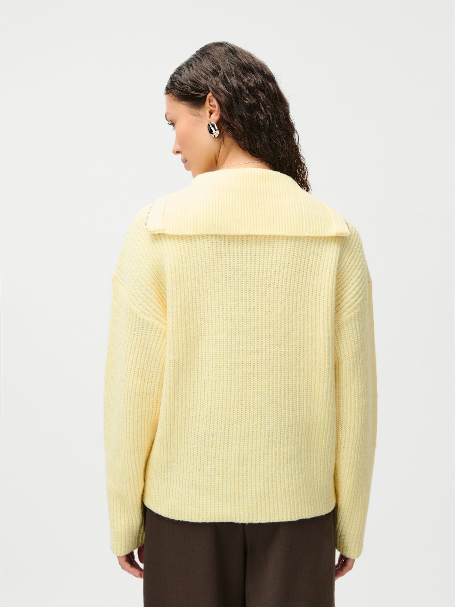 Eine Frau mit gewellten braunen Haaren trägt den LeGer x Purelei Lea Jumper von LeGer by Lena Gercke, einen hellgelben Rippstrickpullover mit lockerer Passform und extralangen Ärmeln, der mit einer braunen Hose kombiniert wird. Sie steht mit dem Rücken zu einem hellen Hintergrund.