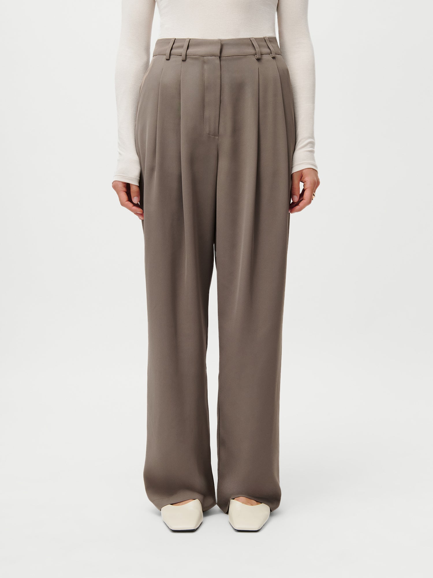 Eine Person (Größe 36, 1,72m) modelliert die LeGer x Purelei - Cara Pants by LeGer by Lena Gercke: eine taupefarbene Hose mit hoher Taille und weitem Bein mit Bundfalten, die mit einem cremefarbenen, langärmeligen Oberteil und cremefarbenen, quadratischen Schuhen vor einem weißen Hintergrund kombiniert wird.