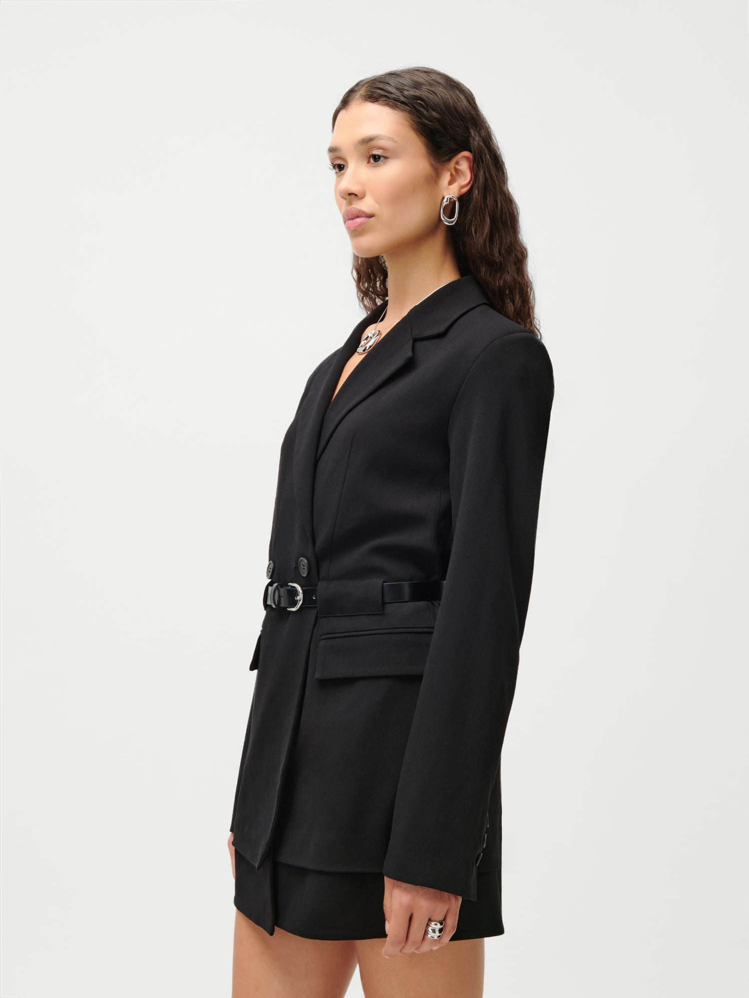 Eine Frau mit welligem braunem Haar trägt das LeGer x Purelei „Jolina“-Kleid von LeGer by Lena Gercke, kombiniert mit silbernen Creolen, einer klobigen Halskette und einem großen Ring. Sie steht vor einem schlichten weißen Hintergrund.