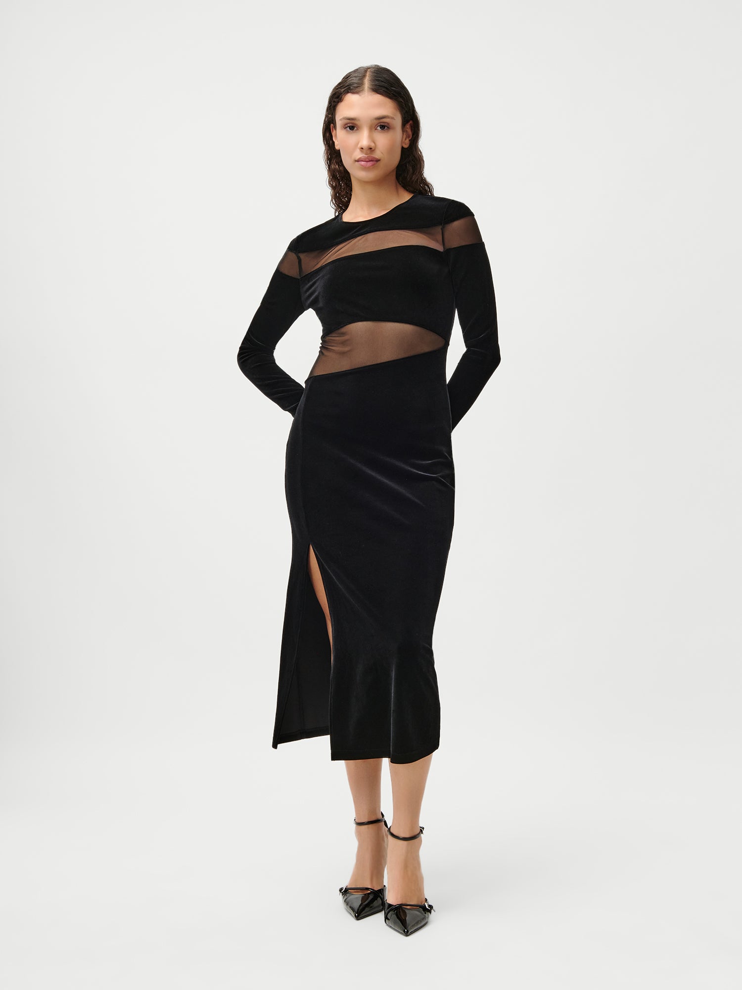 Eine Frau posiert vor einem schlichten weißen Hintergrund und trägt das Fray Dress von LeGer by Lena Gercke, ein elegantes schwarzes langärmeliges Kleid mit transparenten Einsätzen und einem oberschenkelhohen Schlitz. Ihr Haar ist locker und gewellt, und sie kombiniert den Look mit schwarzen spitzen Absätzen mit Knöchelriemen.