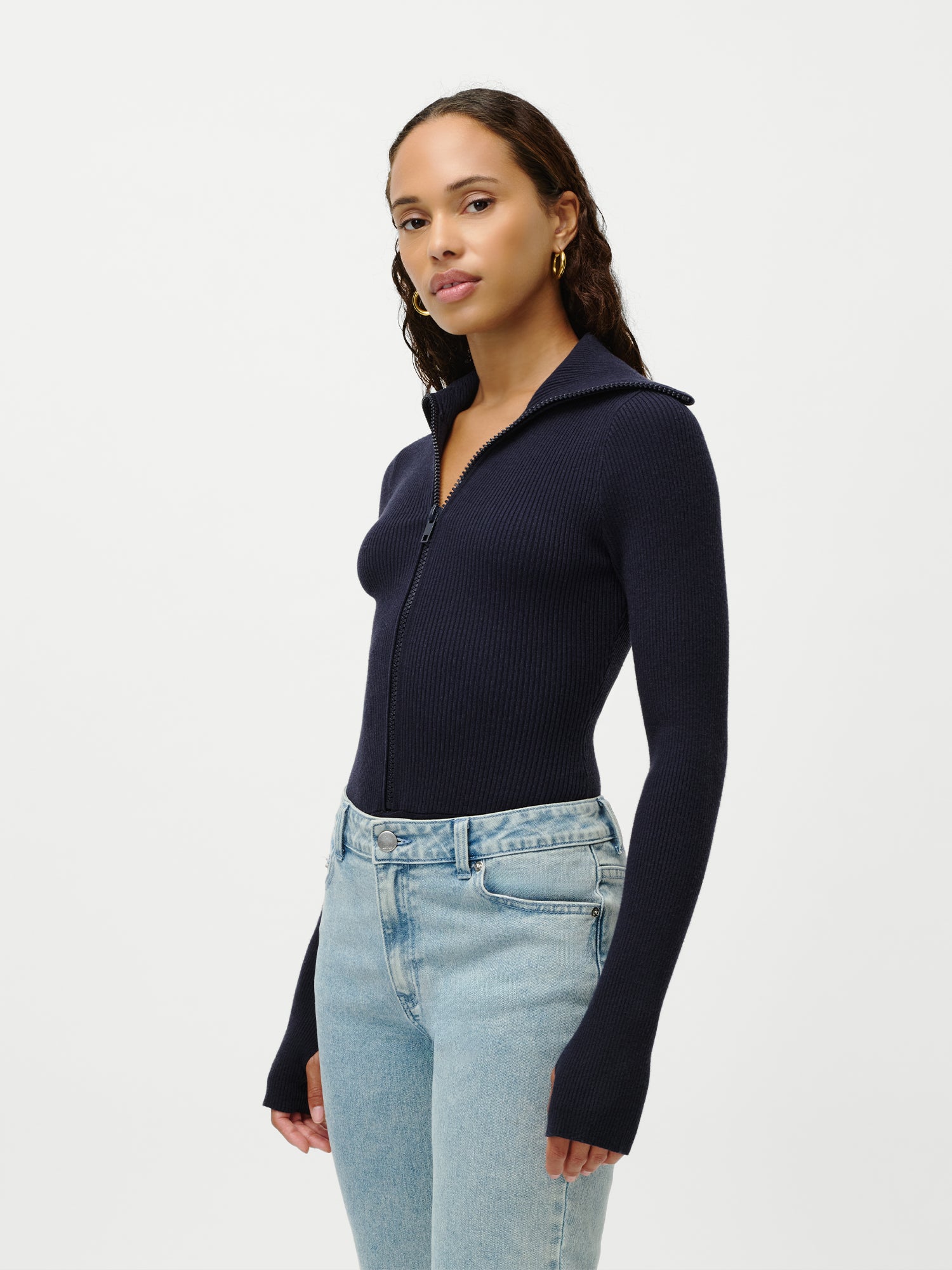 Eine Frau mit langen, gewellten Haaren und goldenen Ohrringen steht vor einem schlichten weißen Hintergrund und trägt den Heidi Body von LeGer by Lena Gercke in Marineblau und hellblaue Jeans mit hoher Taille.