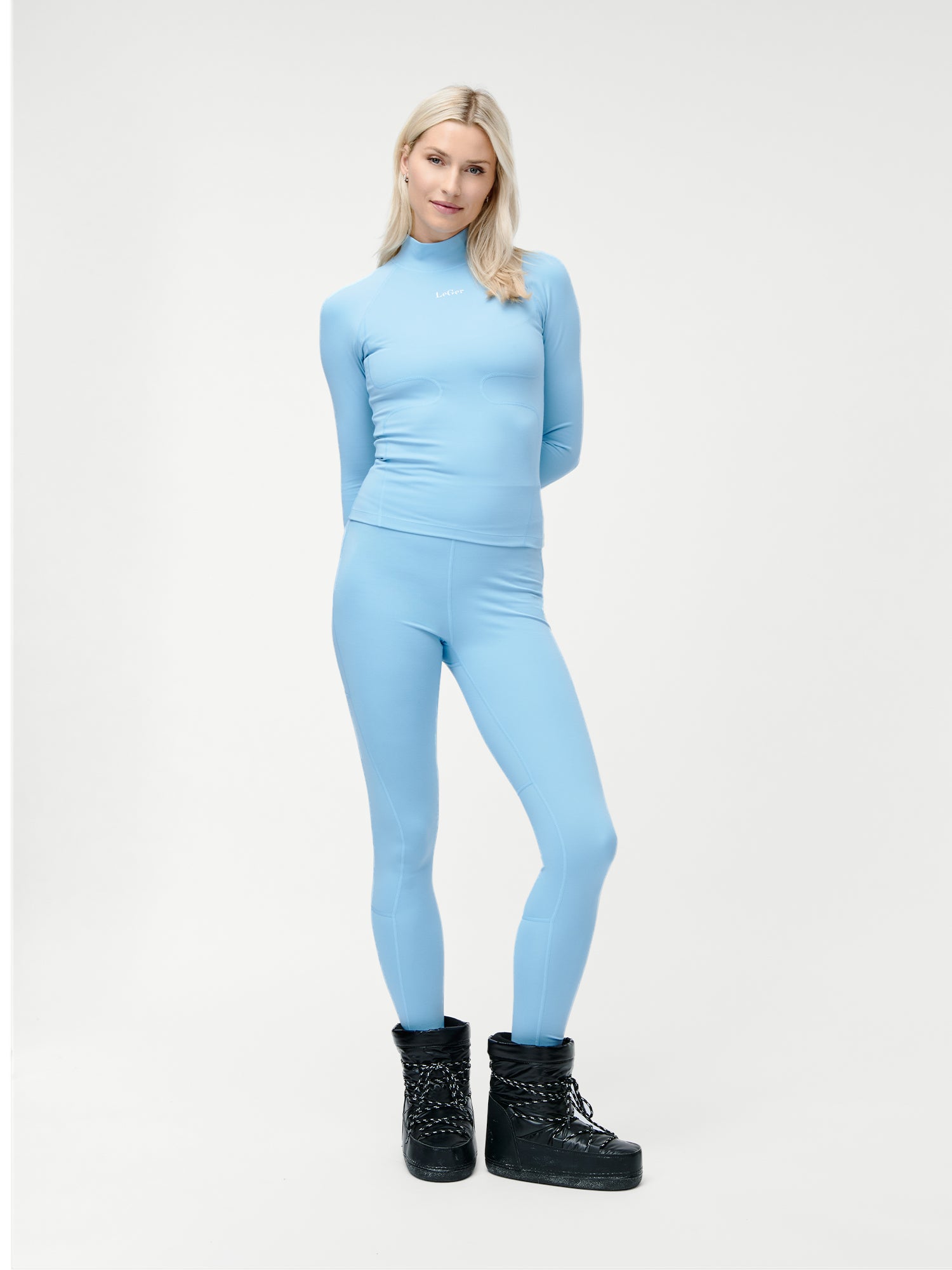Eine Frau mit glattem blondem Haar steht vor einem weißen Hintergrund und trägt die Thalke Pants von LeGer by Lena Gercke mit einem passenden hellblauen, langärmeligen Oberteil und schwarzen Winterstiefeln. Ihr Stil verbindet modernen Komfort mit zeitloser Eleganz.