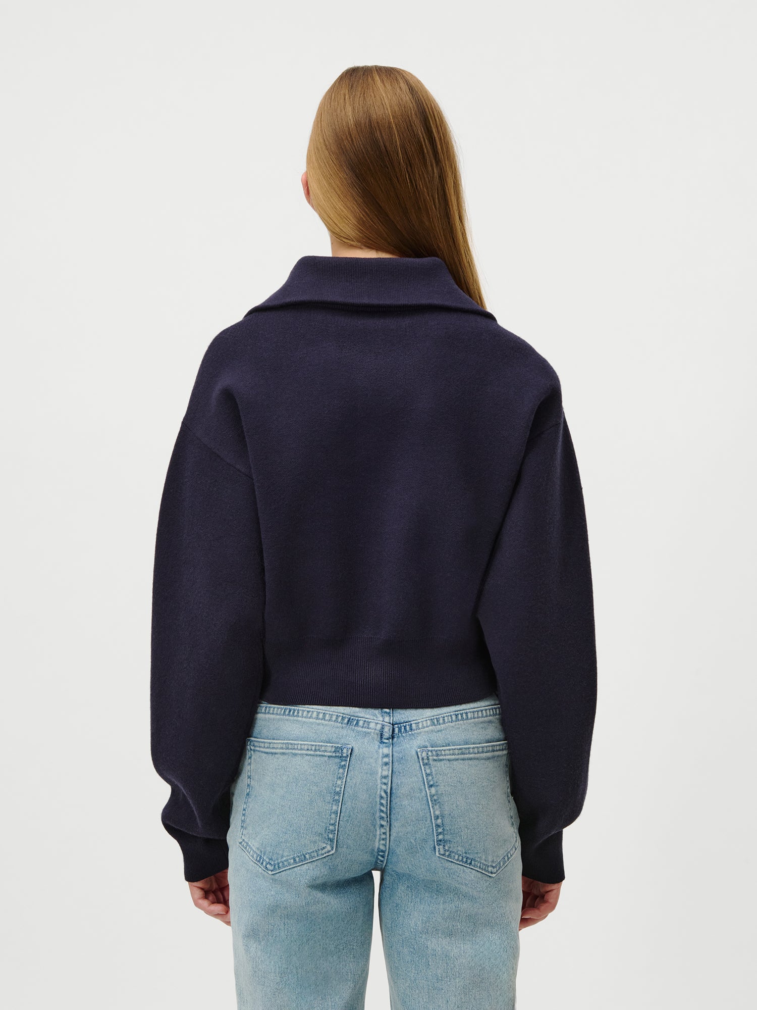 Eine Person mit langem, glattem hellbraunem Haar steht mit dem Rücken zur Kamera und trägt den LeGer by Lena Gercke Jaden Jumper in dark navy mit extra langen Ärmeln und breitem Kragen, gepaart mit einer hellblauen High-Waisted-Jeans der Größe 36 vor einem weißen Hintergrund.
