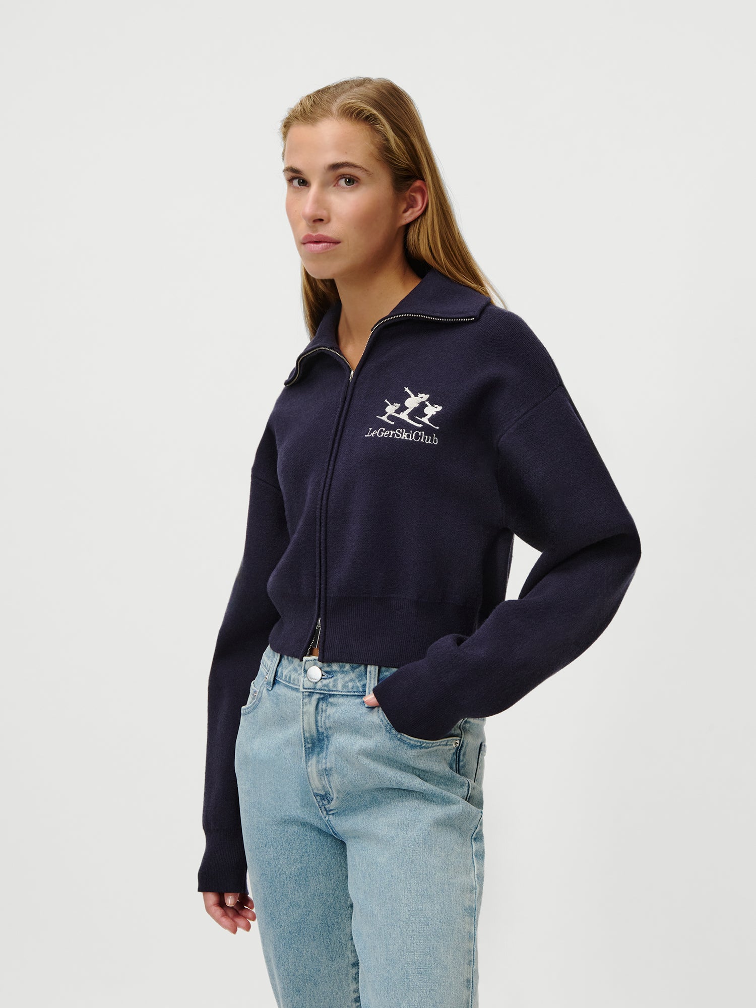 Eine Frau mit langen, hellbraunen Haaren trägt den Jaden Jumper von LeGer by Lena Gercke - einen marineblauen Pullover mit Reißverschluss, extralangen Ärmeln und weißem Loro Piana Ski Club"-Logo - zusammen mit einer hellblauen Jeans in Größe 36 vor einem schlichten weißen Hintergrund.