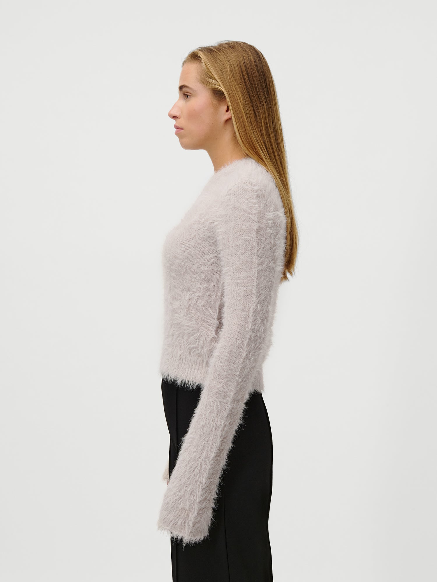 Eine Frau mit langen, glatten Haaren steht im Profil vor einem schlichten weißen Hintergrund. Sie trägt den Pullover Sharon in Größe 36 von LeGer by Lena Gercke und eine schwarze Hose.