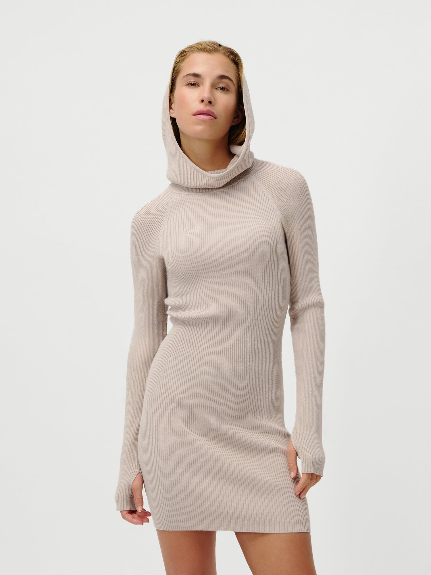 Eine Frau trägt das LeGer by Lena Gercke Mathilde Dress, ein schmal geschnittenes, hellbeiges Rippstrickkleid mit extralangen Ärmeln und Daumenlöchern. Sie steht selbstbewusst vor einem weißen Hintergrund und hält die Hände an den Seiten.
