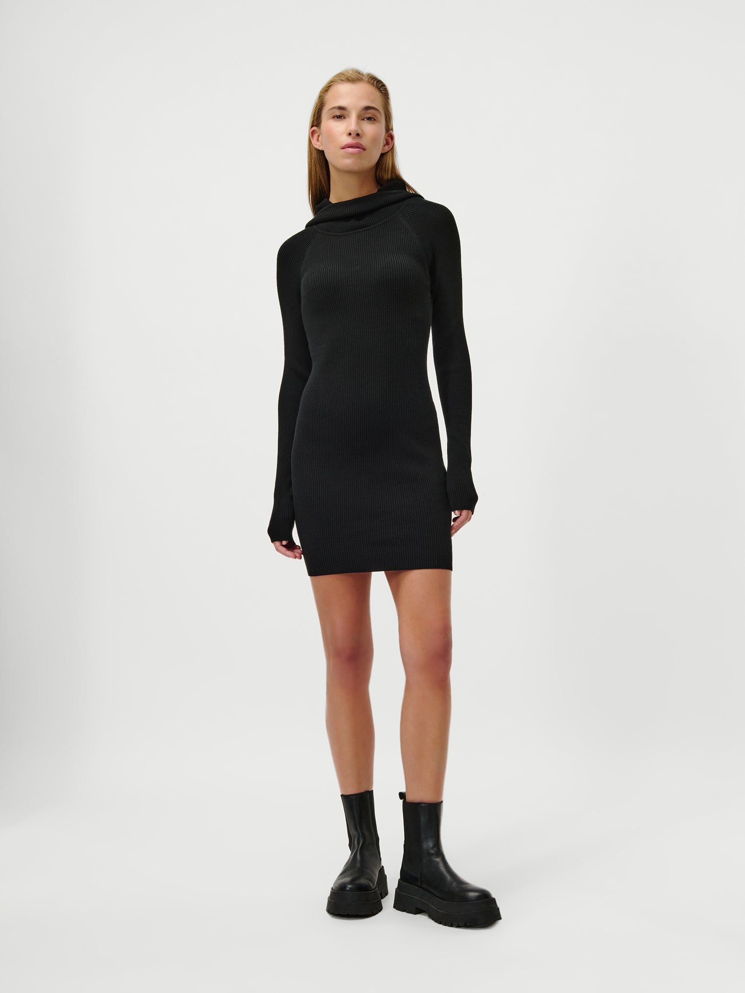 Eine Person steht vor einem schlichten weißen Hintergrund und trägt das Mathilde Dress von LeGer by Lena Gercke, ein eng anliegendes, langärmeliges Minikleid mit extralangen Ärmeln und schwarzen Ankle Boots. Sie hat glattes, schulterlanges Haar und einen neutralen Gesichtsausdruck.