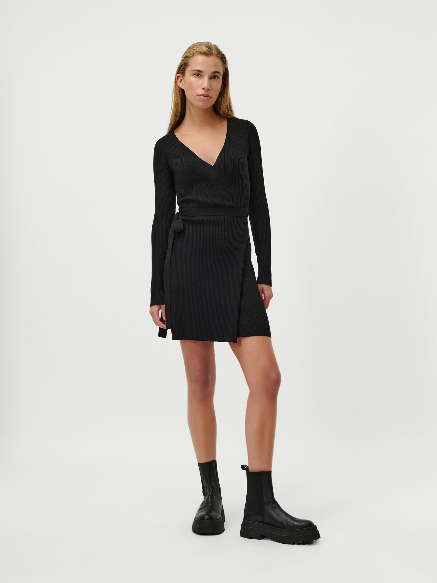 Eine Frau mit glattem hellbraunem Haar und neutralem Gesichtsausdruck trägt das LeGer by Lena Gercke Effie Dress, ein tailliertes schwarzes langärmeliges Wickelkleid mit V-Ausschnitt, gepaart mit schwarzen Ankle Boots, vor einem schlichten weißen Hintergrund.