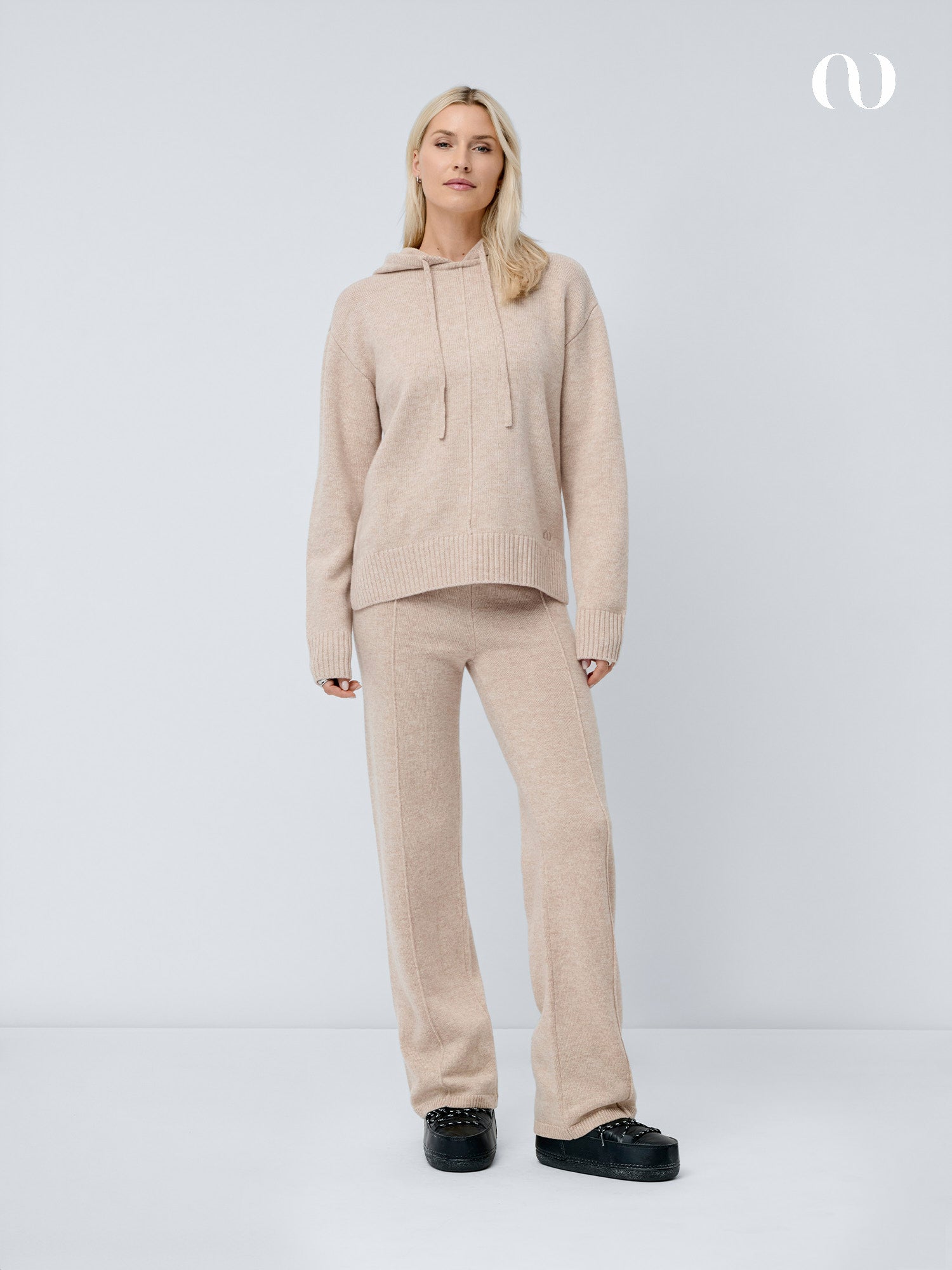 Eine Frau mit blonden Haaren steht vor einem unifarbenen Hintergrund und trägt die beigefarbene Godja Pants von LeGer by Lena Gercke in einem gemütlichen Pull-on-Design mit einer passenden Strickkapuzenjacke und schwarzen Schuhen. In der rechten oberen Ecke ist ein weißes Logo zu sehen.