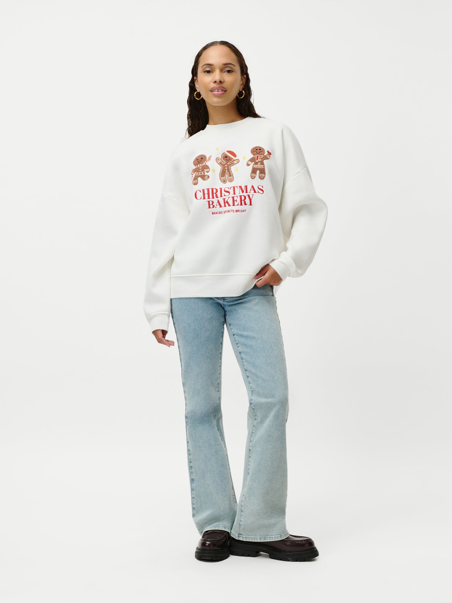 Vor einem weißen Hintergrund steht eine Frau mit langen, welligen Haaren. Sie trägt das Sweatshirt „Daphne“ aus der Weihnachtskollektion von LeGer by Lena Gercke mit Lebkuchenfiguren, hellblaue Jeans in normaler Länge und schwarze Schuhe und wirkt dabei ganz entspannt.